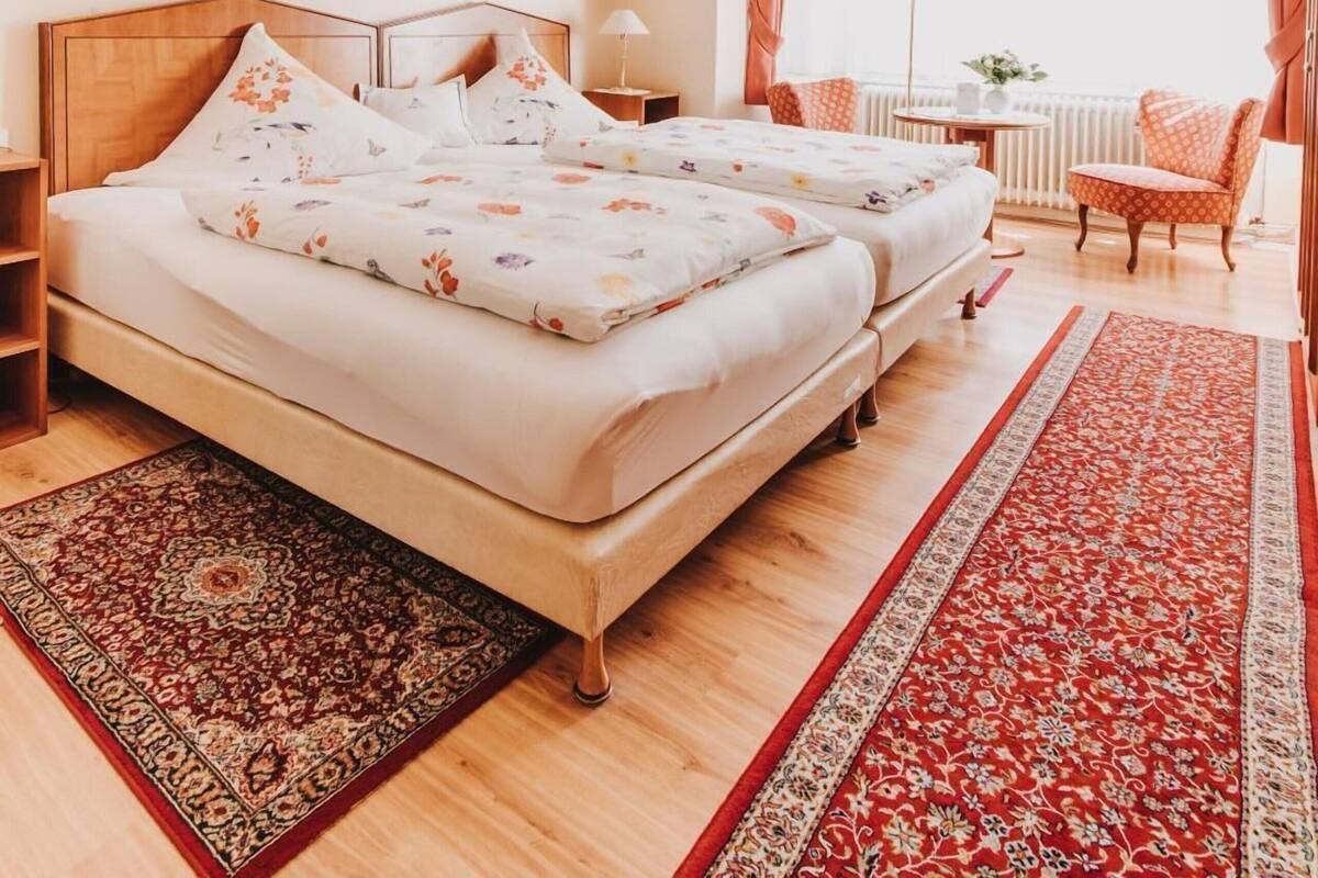 A bedroom with a large bed, a chandelier, a window with curtains, a small table, and a chair.