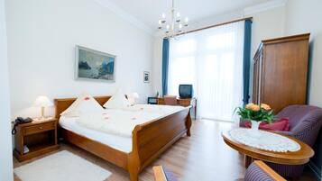 A hotel room with a wooden bed, a desk with a computer, a chair, a small table with flowers, and a painting on the wall.