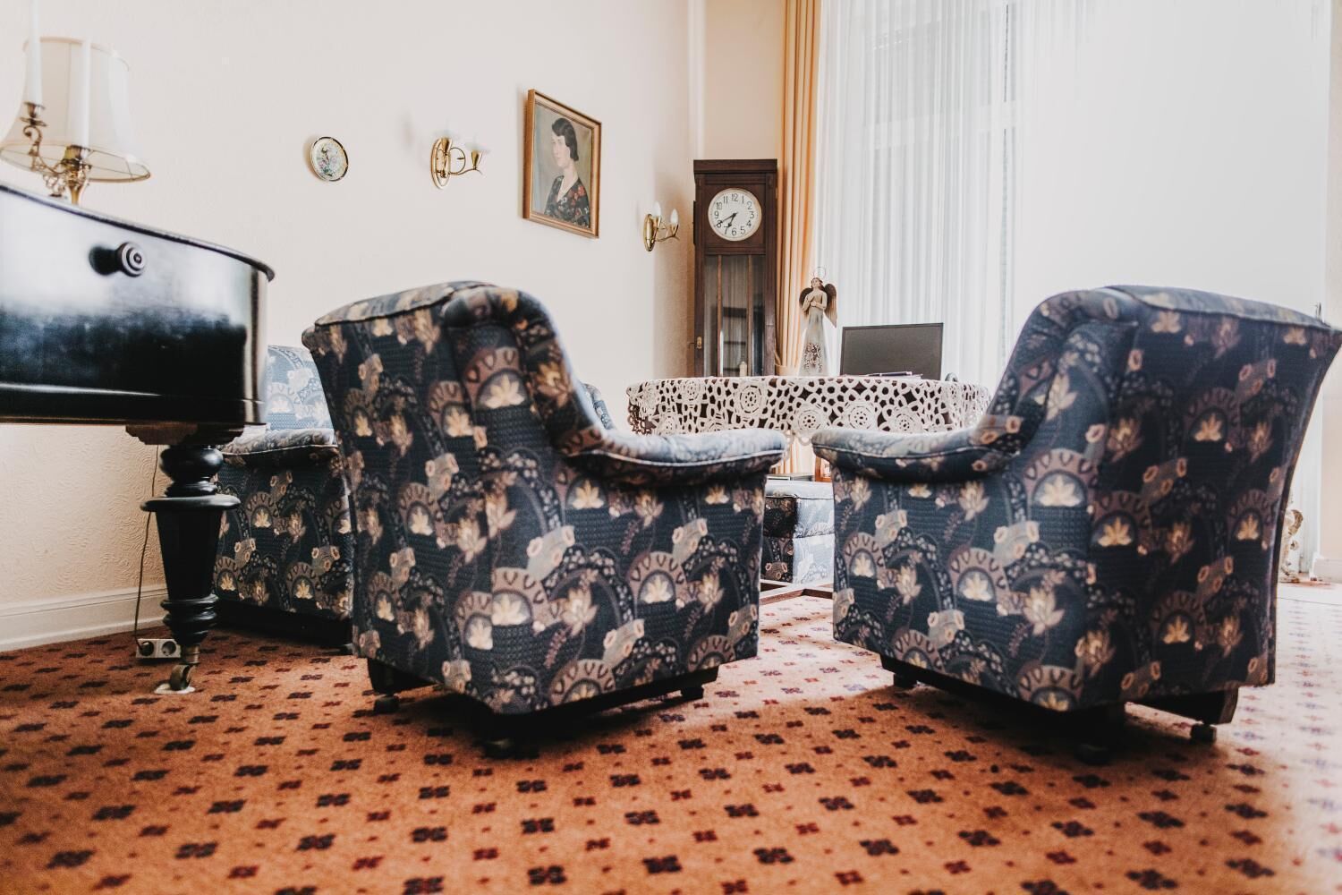 A room with a piano, two patterned armchairs, a lace table, a clock, and a framed picture on the wall.