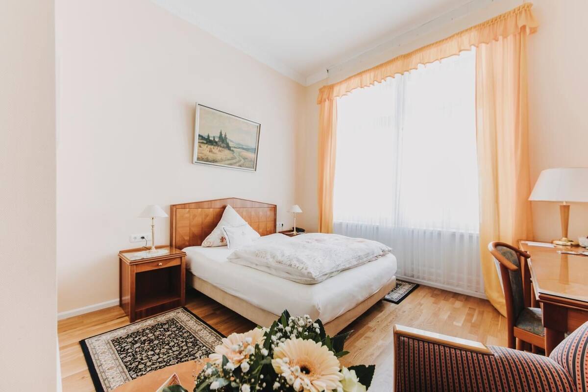 A hotel room with a bed, two bedside tables, a chair, a desk, a window with curtains, and a painting on the wall.