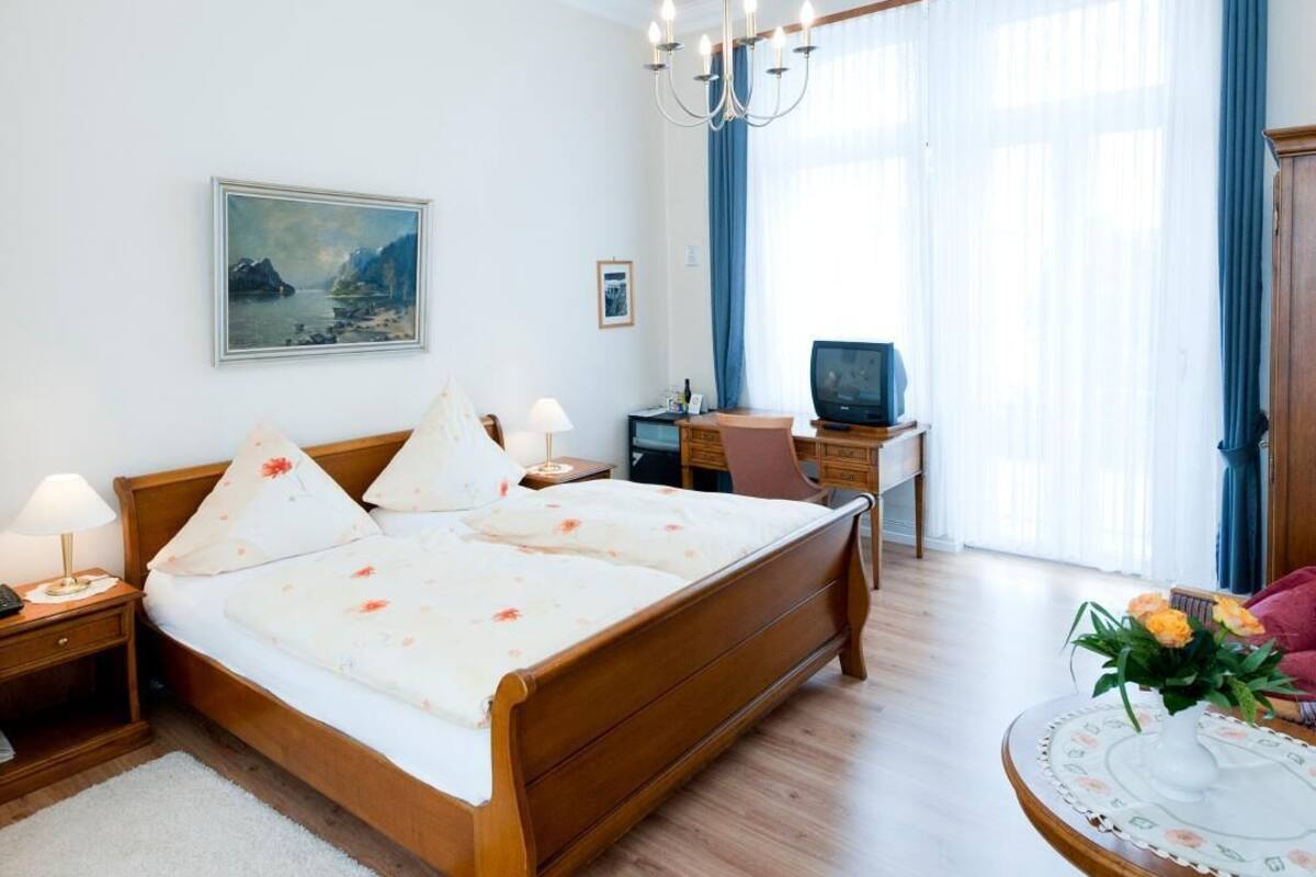 A hotel room with a wooden bed, a desk with a computer, a chair, a small table with flowers, and a painting on the wall.