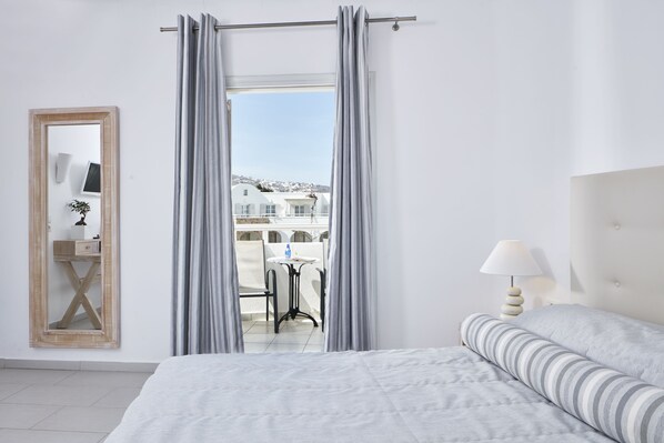 A hotel room with a bed, a bedside table with a lamp, a mirror, and a view of the outside balcony. - Aegean Plaza Hotel (Santorini)