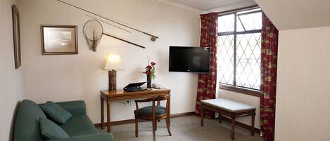 A hotel room with a green sofa, a small wooden table with a lamp, a television, and a window with curtains.