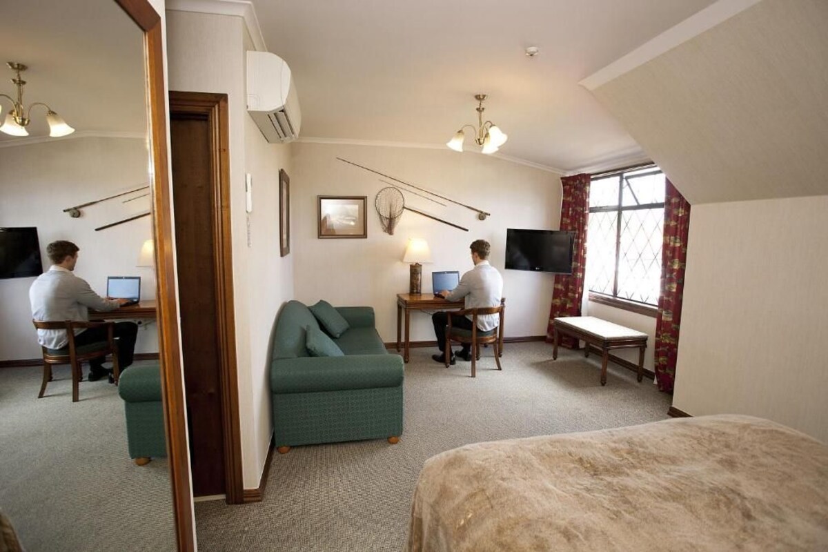 A hotel room with two workstations, a green sofa, a bed, and a large window with curtains.