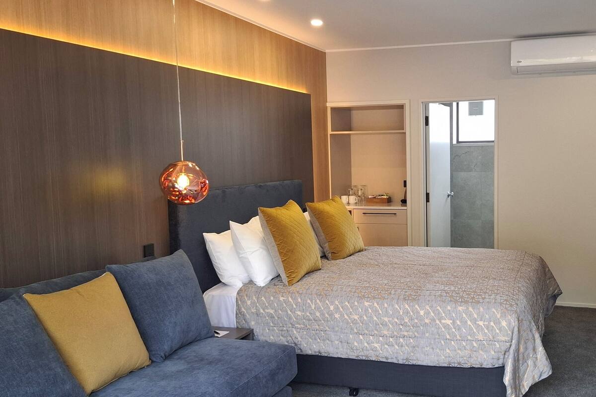 A modern hotel room with a bed, a sofa, a TV, and a bathroom visible through an open door.