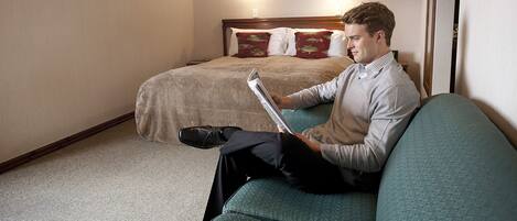 A man sitting on a teal sofa in a hotel room, reading a magazine. The room has a wooden headboard bed with red and green pillows, a carpeted floor, and a small table with a lamp.