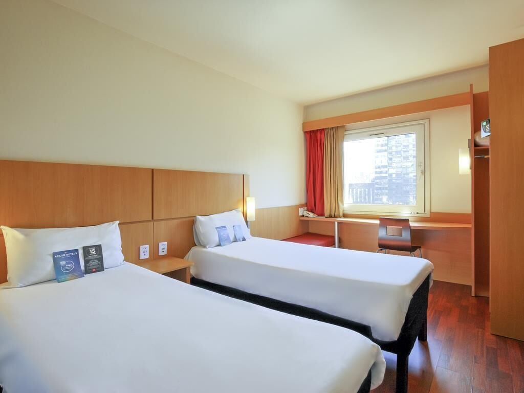 A hotel room with two beds, a desk, a window with curtains, and a view of the city.