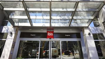Entrada de un hotel con marquesina de vidrio, señalización de 'ibis' y un contenedor de basura con la etiqueta 'RESIDIOS'.