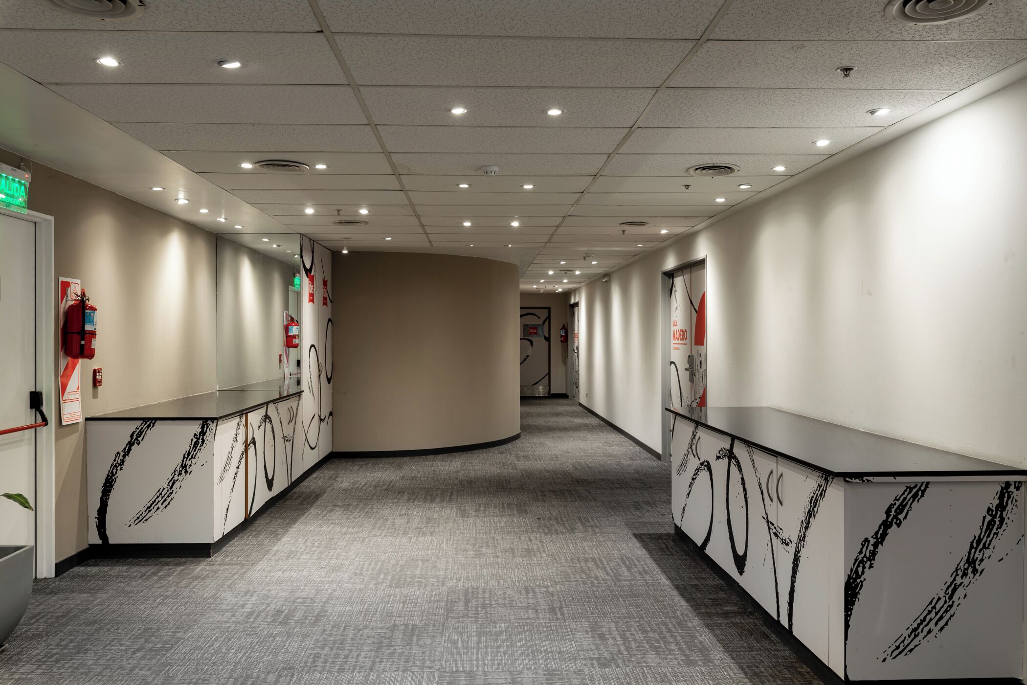 A hallway with a curved reception desk, fire extinguishers, and emergency exit signs.