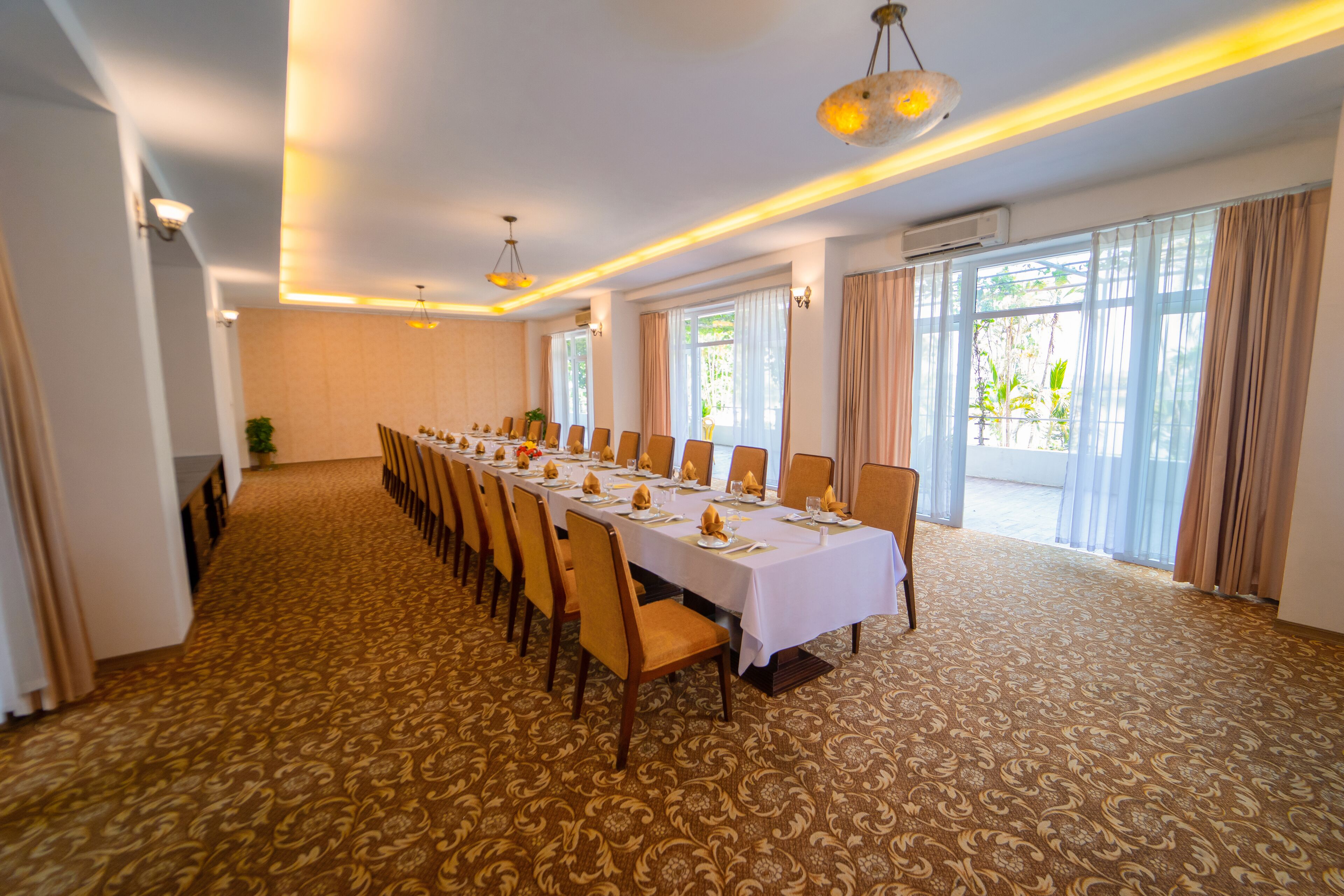 A long dining table set for a meal in a well-lit room with patterned carpet, large windows, and curtains.