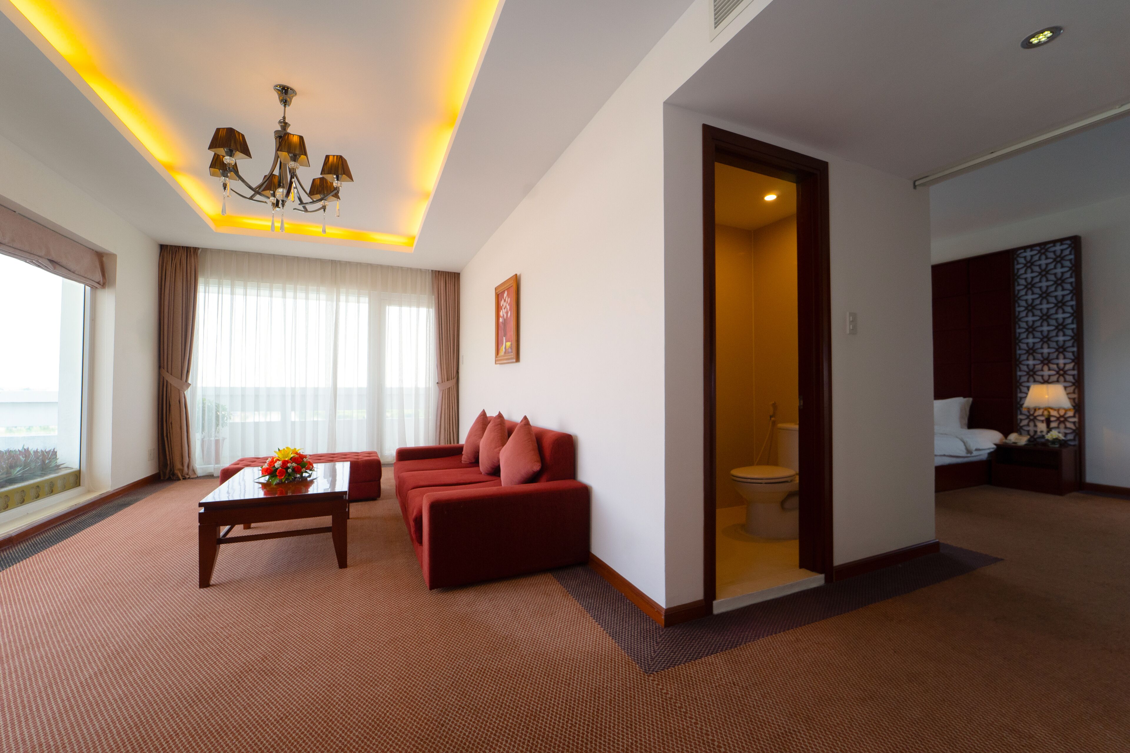Une chambre d’hôtel avec un canapé rouge, une table basse et un lustre. On peut apercevoir la salle de bain à travers une porte ouverte.