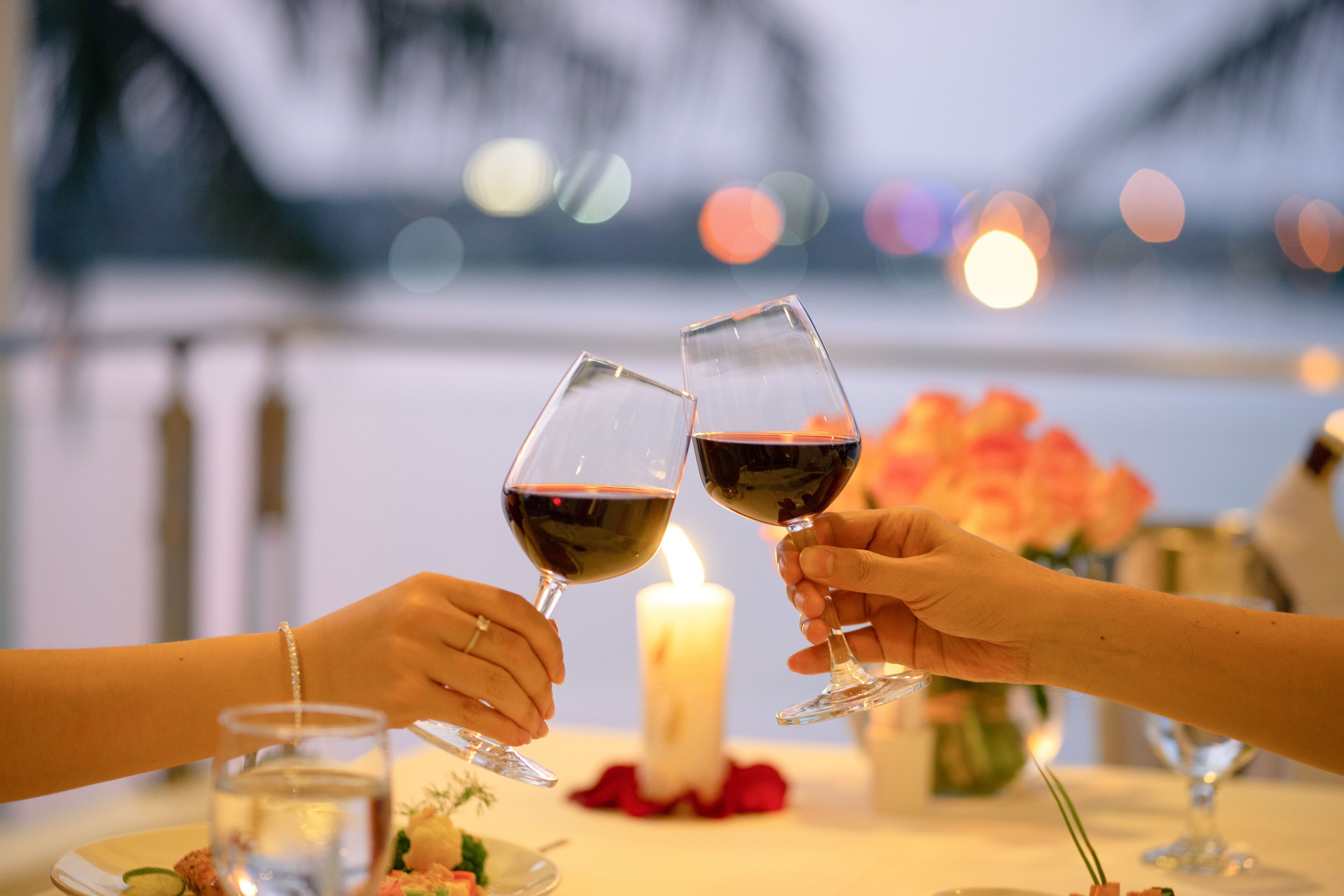 Two people toasting with wine glasses at a table with a candle and flowers, overlooking a body of water.