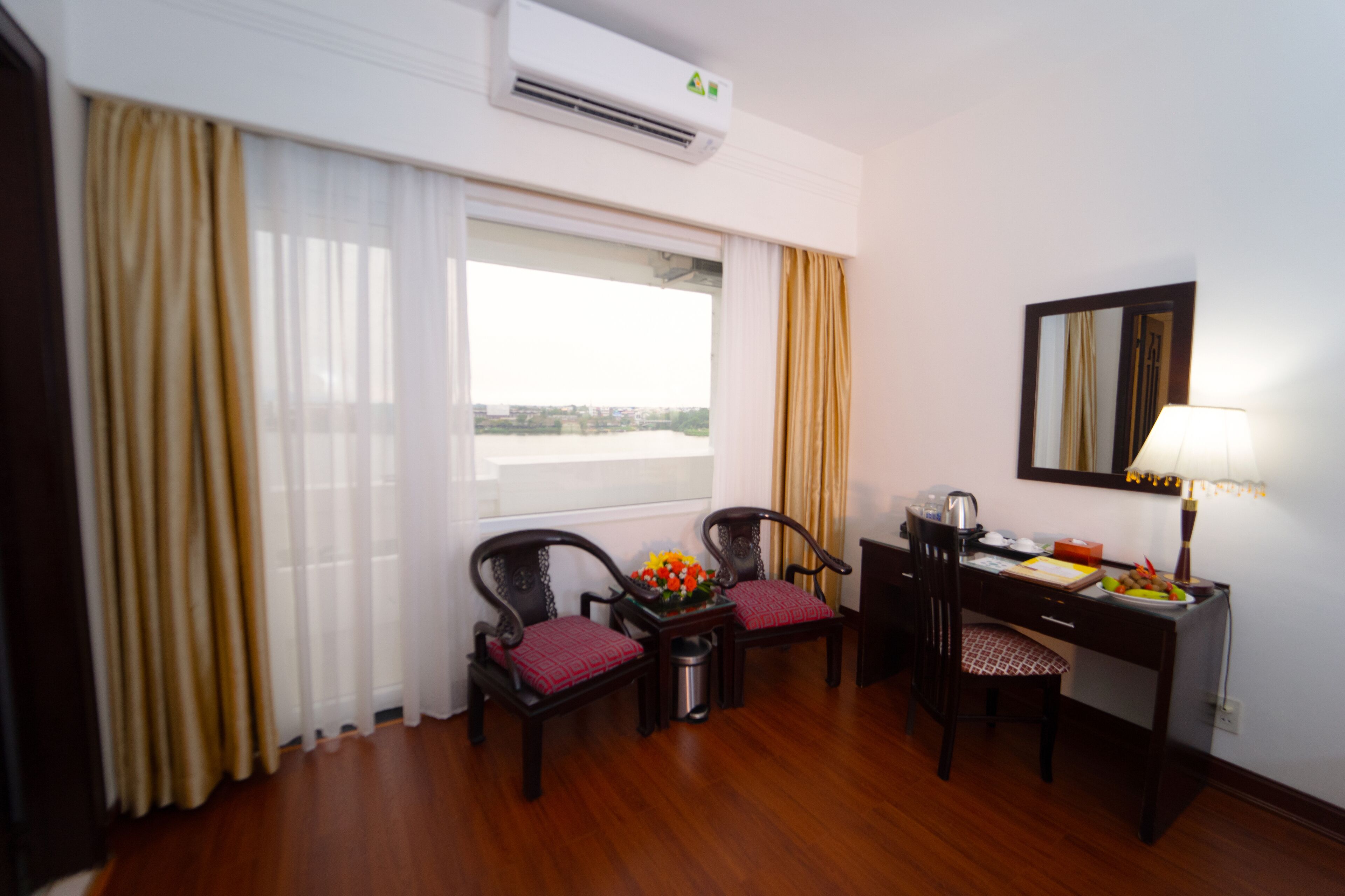 A hotel room with a wooden floor, a desk with a lamp, two chairs, a small trash bin, and a window with curtains.