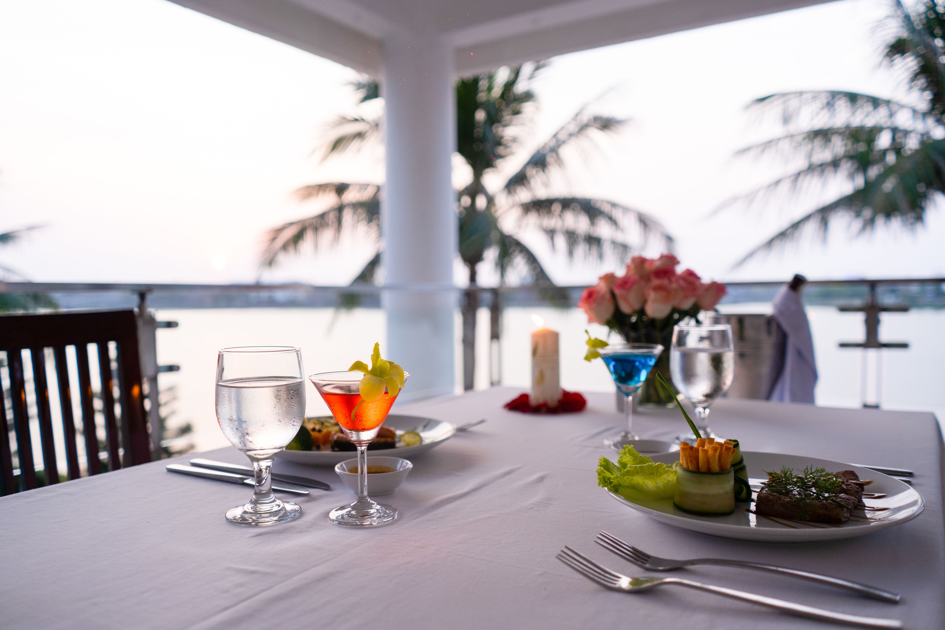 A dining table set with a glass of water, a cocktail, and plates of food, overlooking a beach with palm trees.