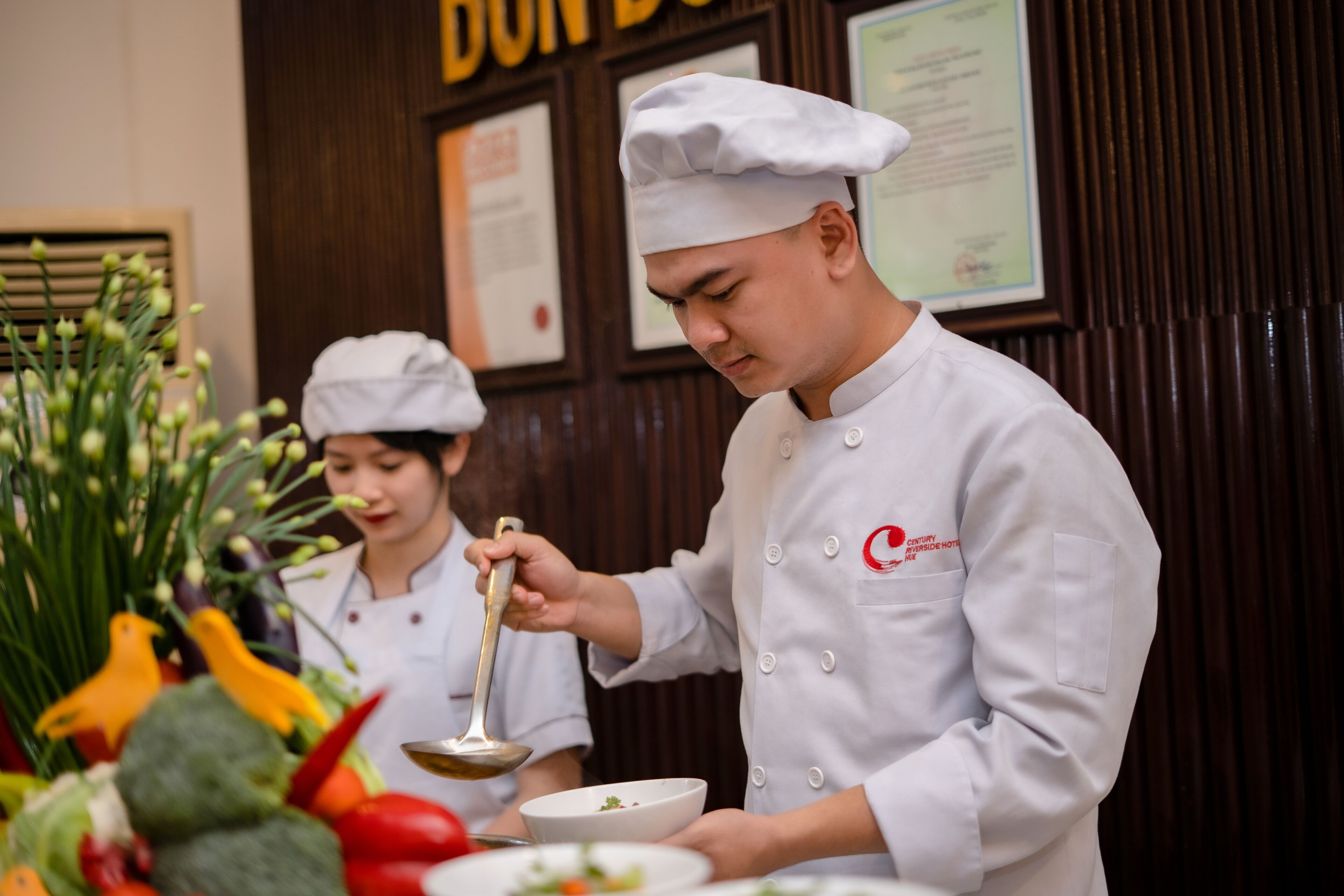 A chef in a white uniform with a red logo, holding a ladle over a bowl, with a variety of vegetables in the foreground.