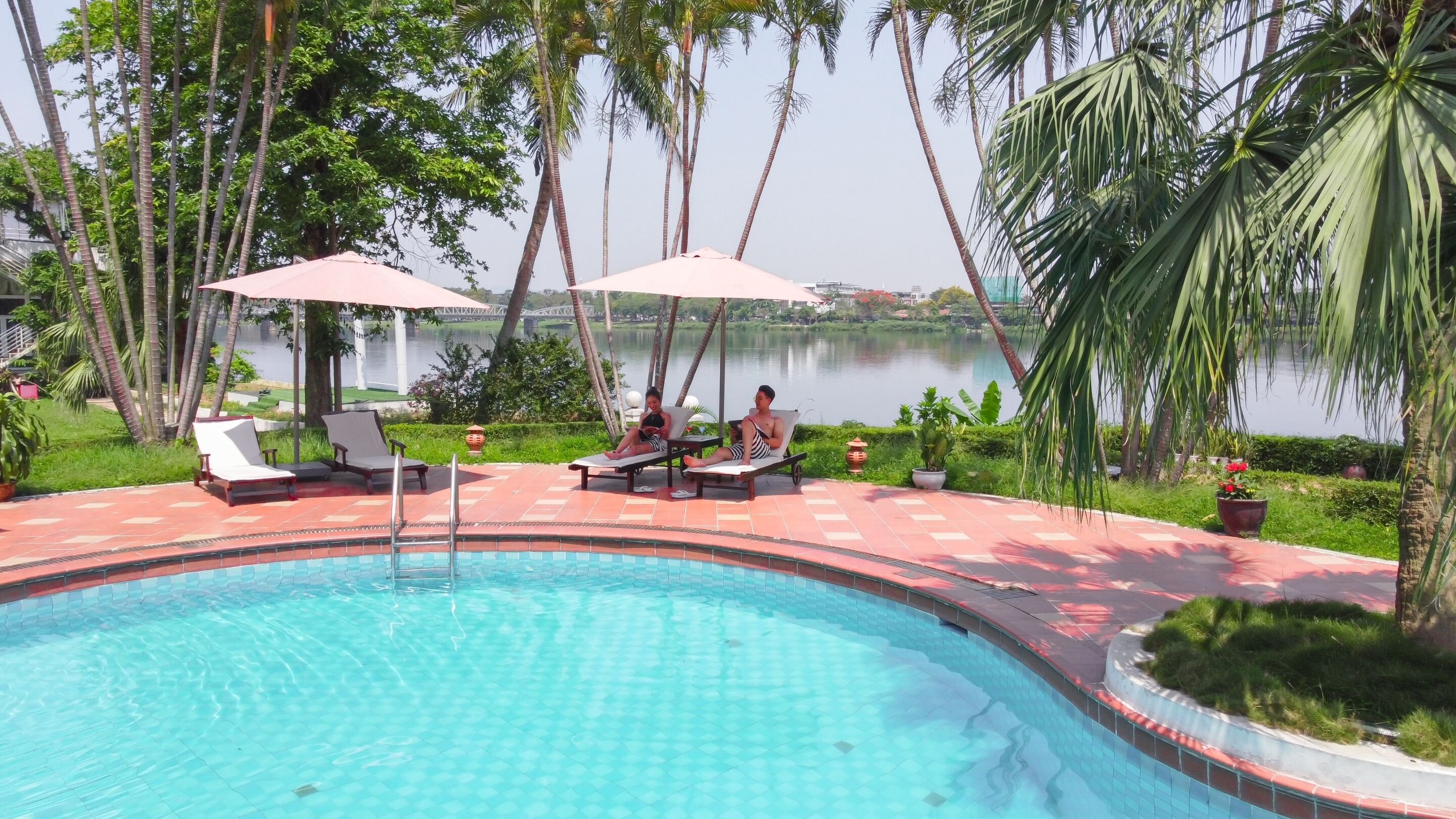 Une piscine entourée de palmiers, avec des chaises longues et des parasols, et offrant une vue sur un lac.