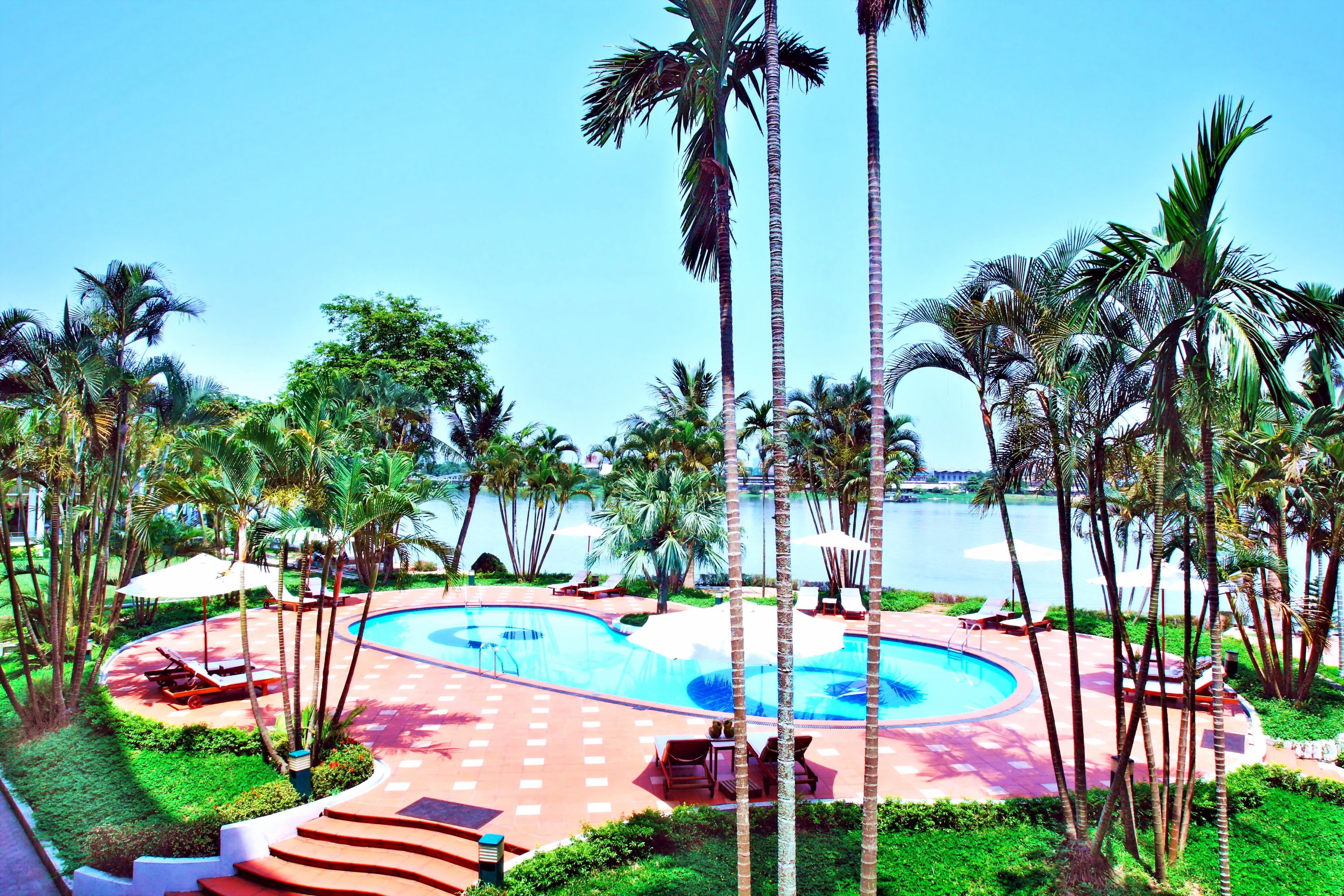 Zona de piscina en un resort, con palmeras, sillas de descanso y vista despejada al agua.