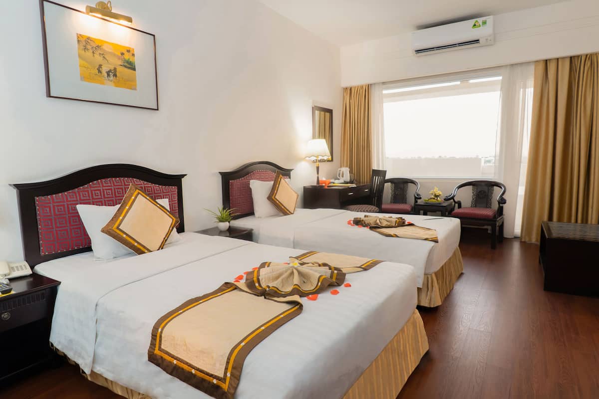 A hotel room with two beds, a desk with a lamp, a chair, a small table with a plant, and an air conditioning unit.