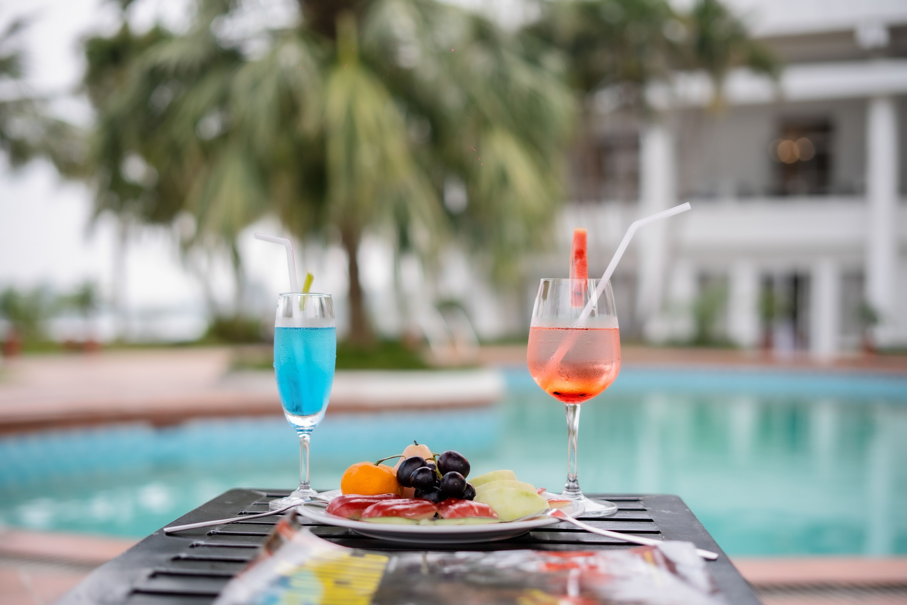 A table with two drinks and a plate of fruit by a poolside.