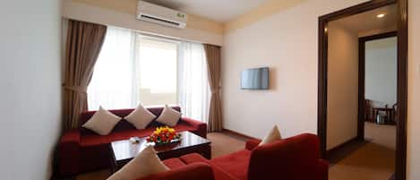 Quarto de hotel com sofá vermelho, mesa de centro com arranjo floral, TV de tela plana e uma grande janela com cortinas.