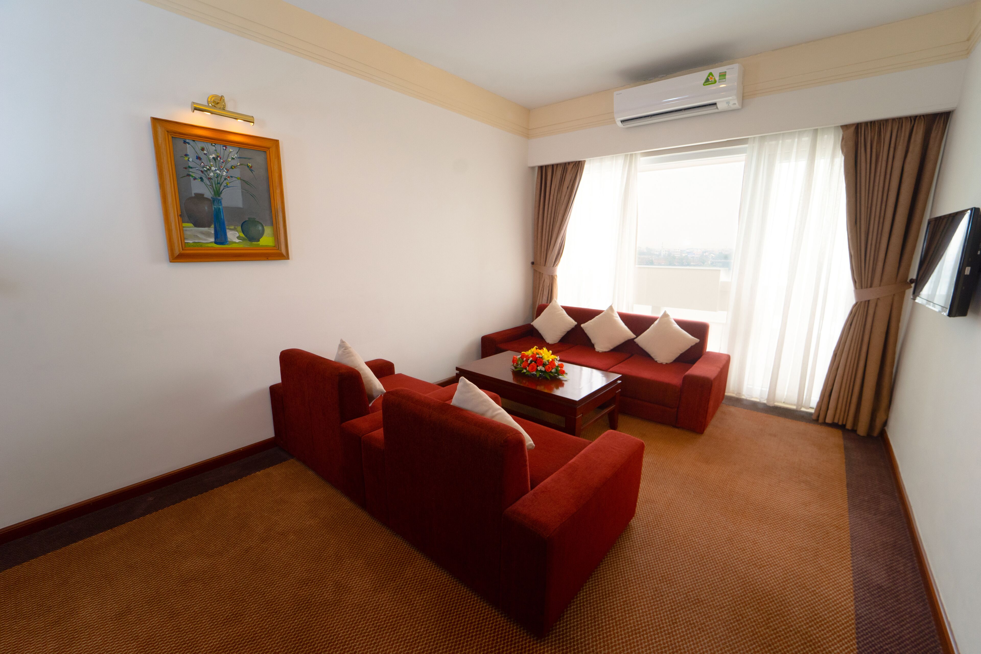 Habitación de hotel con un sofá rojo, una mesita de centro con flores, un cuadro en la pared y un equipo de aire acondicionado.