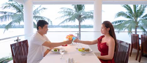 Una mesa de comedor con mantel blanco, dos personas brindando y vistas a palmeras y agua.