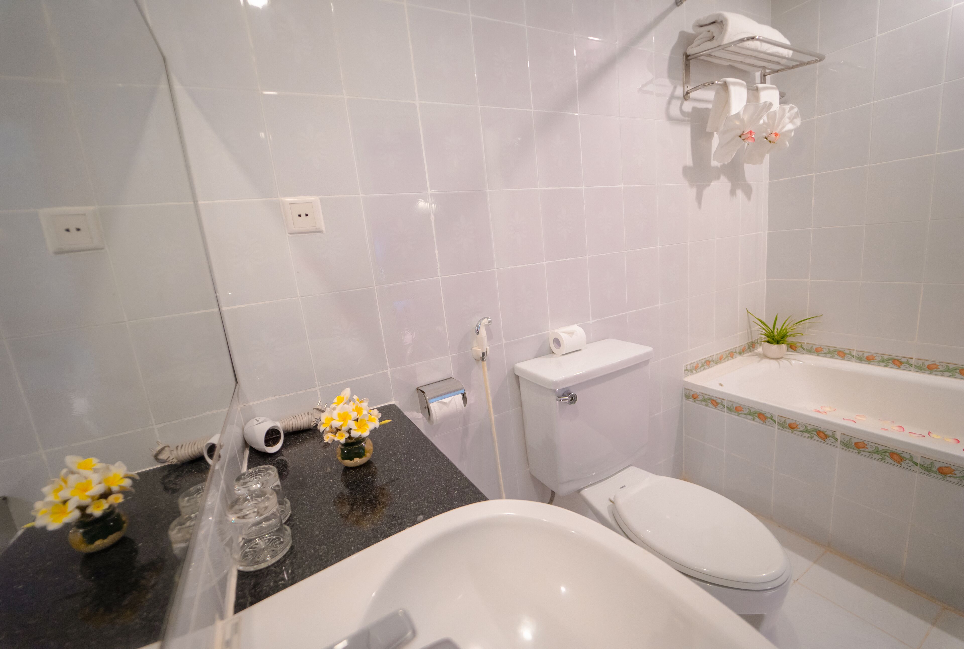 A bathroom with a toilet, sink, and bathtub. There are flowers on the counter and a plant on the bathtub edge.