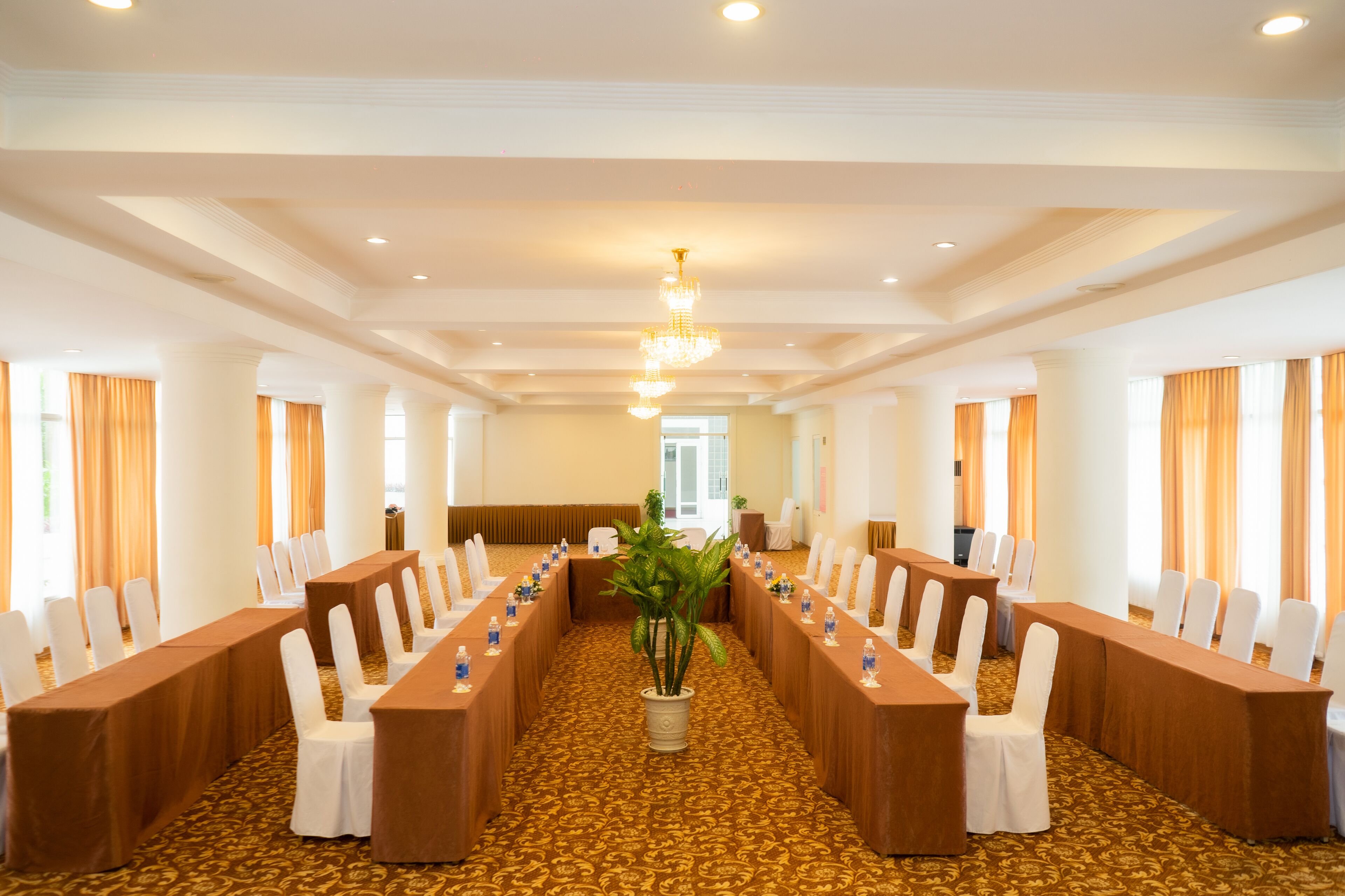 A conference room with long tables and chairs, a chandelier, and a plant in the center.
