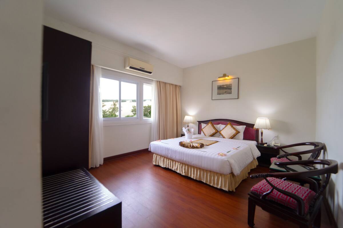A hotel room with a bed, bedside tables, a chair, a window with curtains, and an air conditioning unit.