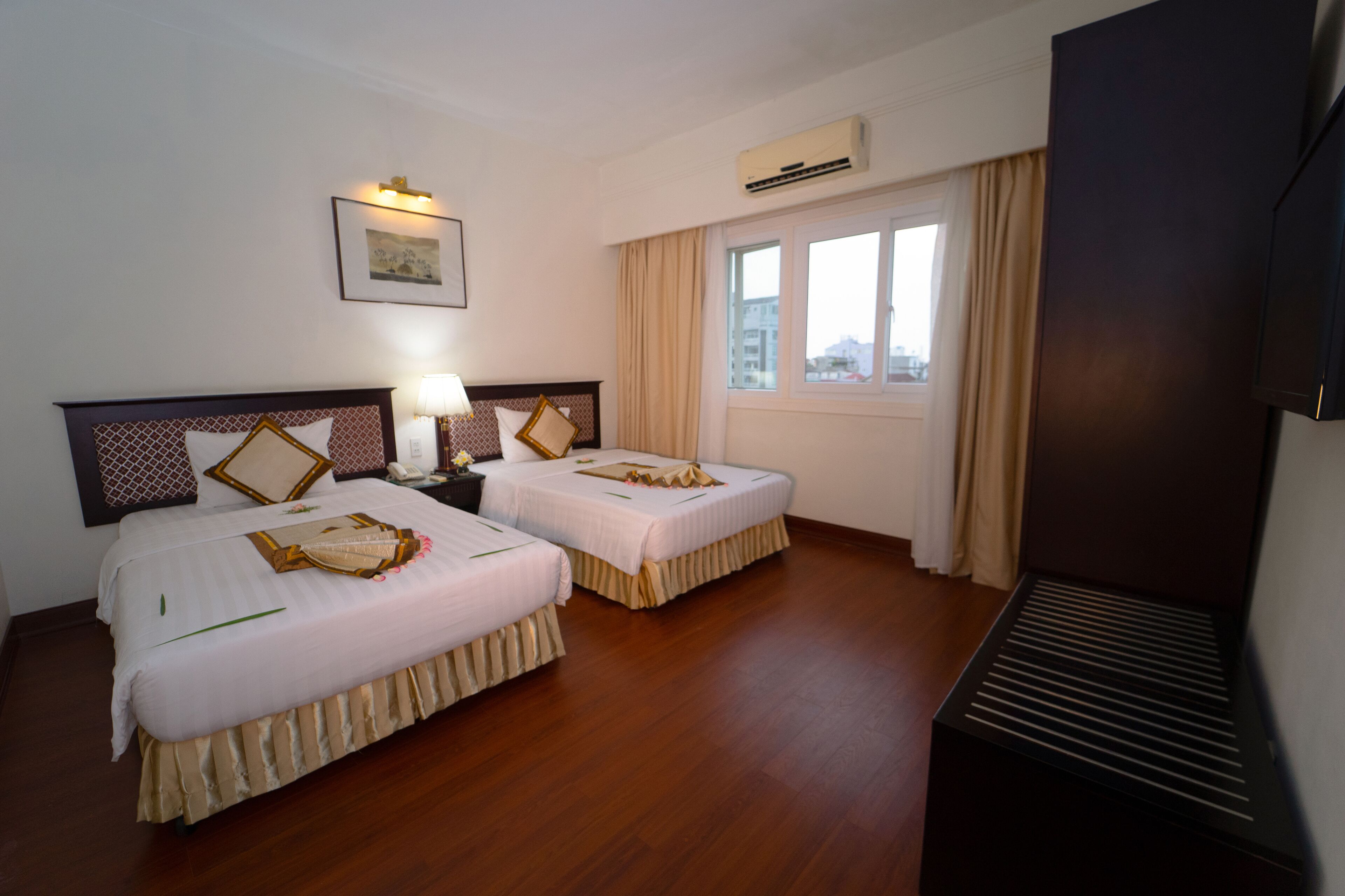A hotel room with two beds, a bedside table with a lamp, an air conditioning unit, a window with curtains, and a television.