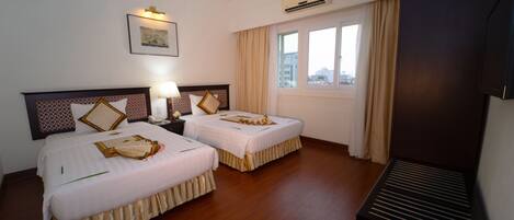 A hotel room with two beds, a bedside table with a lamp, an air conditioning unit, a window with curtains, and a television.