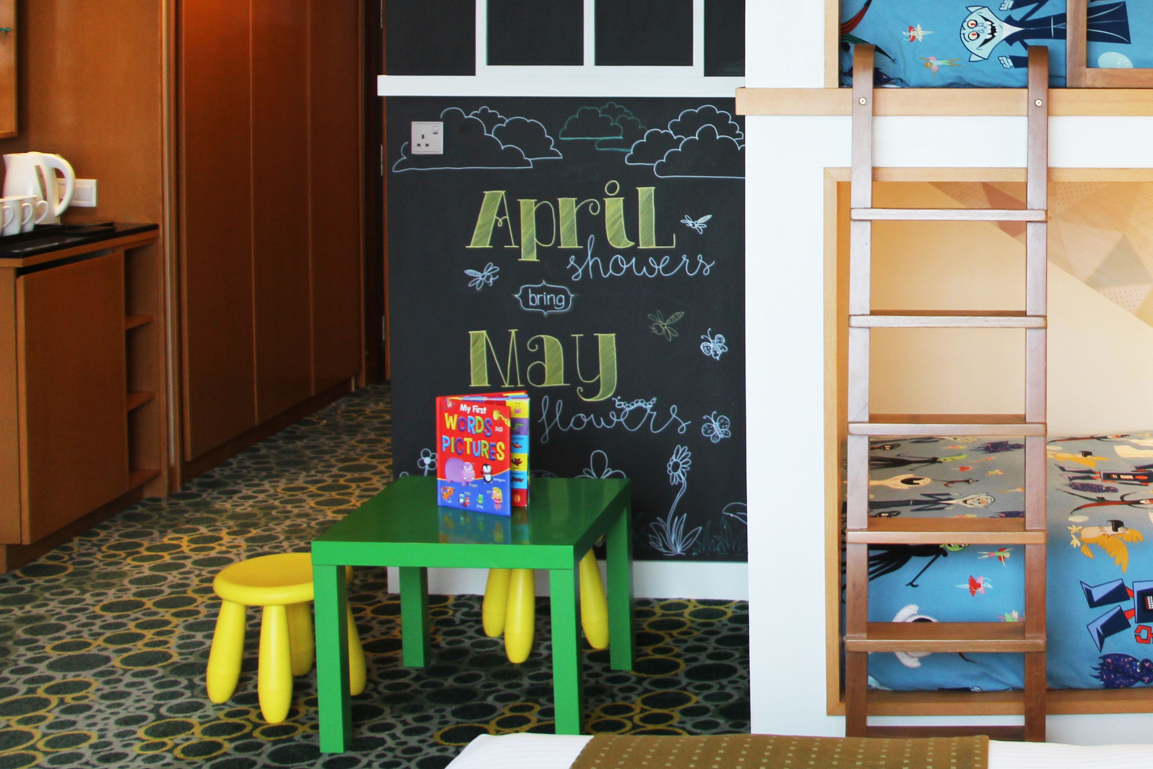 Una habitación infantil con un muro de pizarra, una mesa verde con taburetes amarillos y una cama litera con ropa de cama estampada.