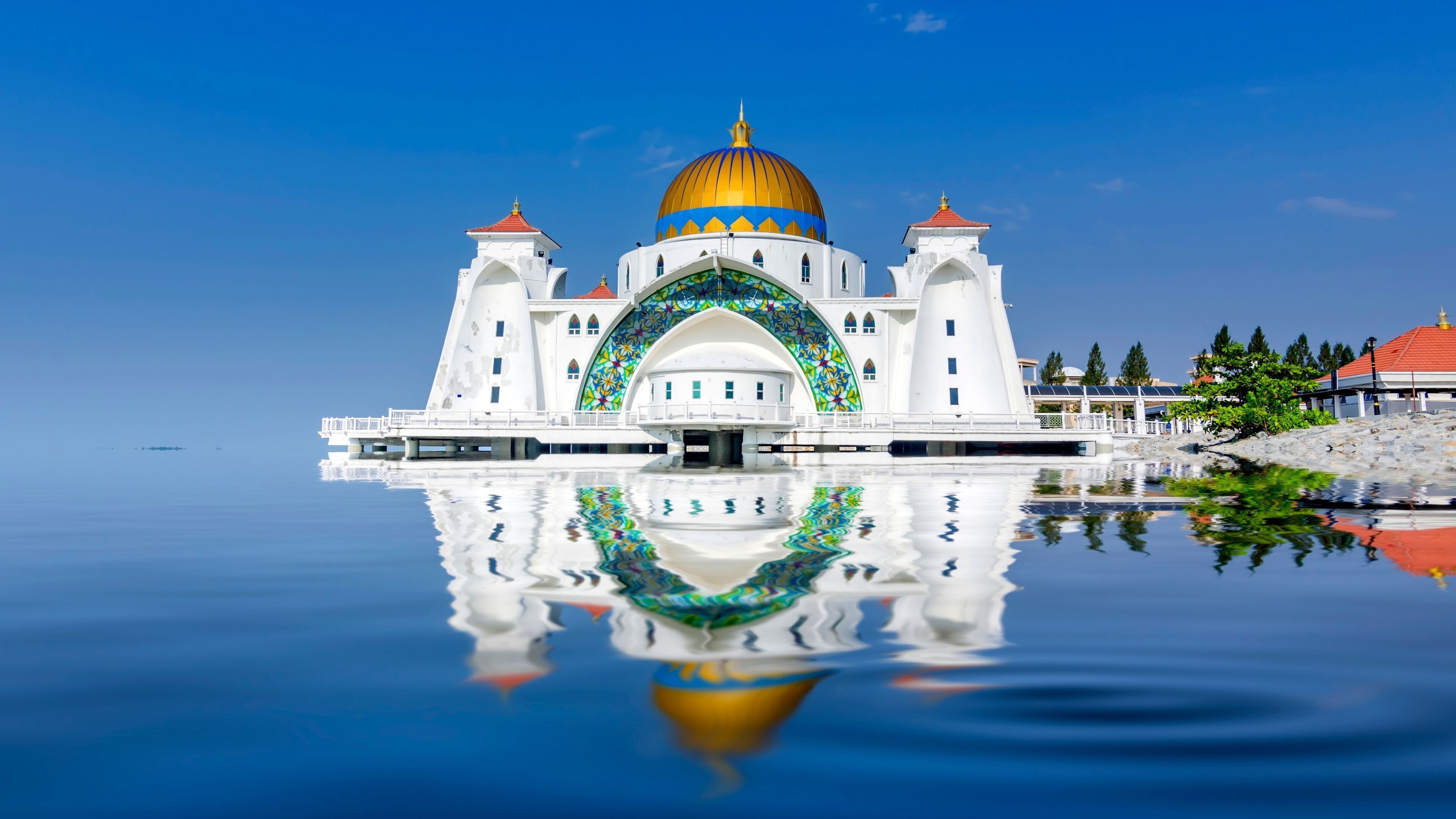 A white building with a golden dome and a large arched entrance, reflecting in calm water.