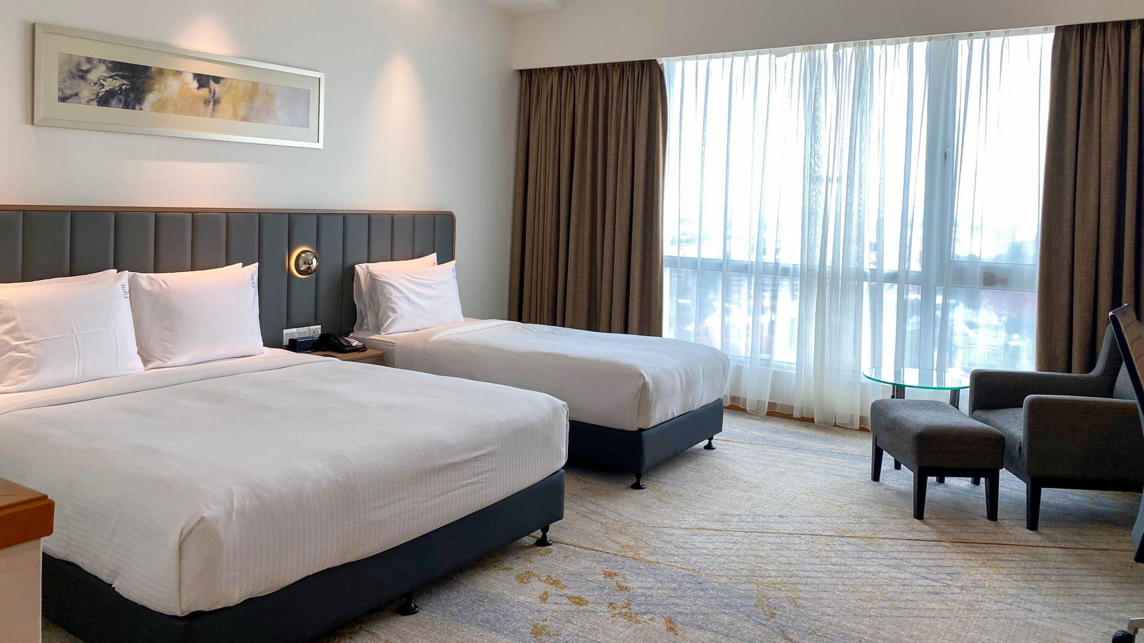 A hotel room with two beds, a desk, a chair, a painting on the wall, and a large window with curtains.