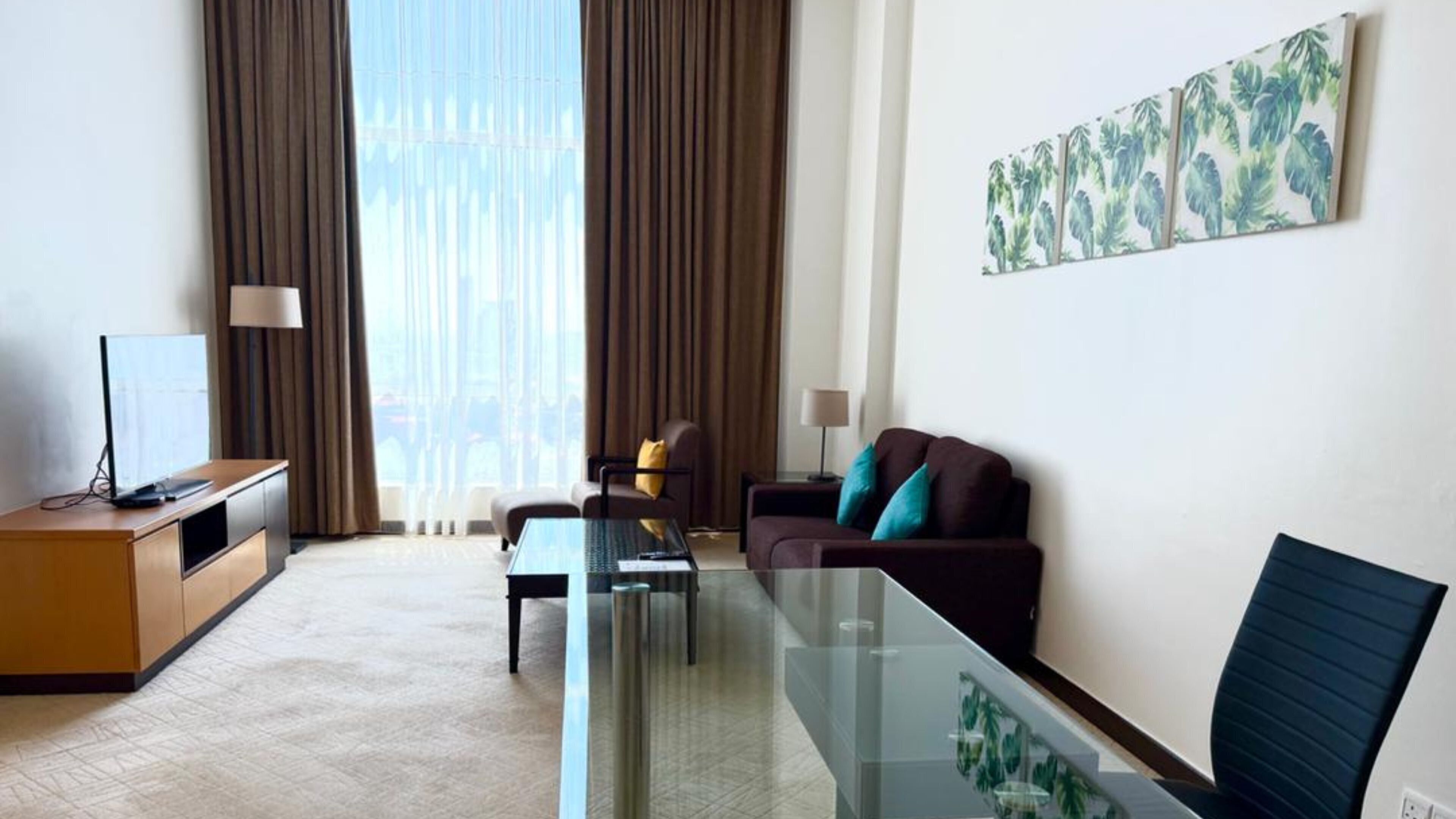 A modern hotel room with a glass dining table, a sofa, a TV on a wooden cabinet, and large windows with curtains.