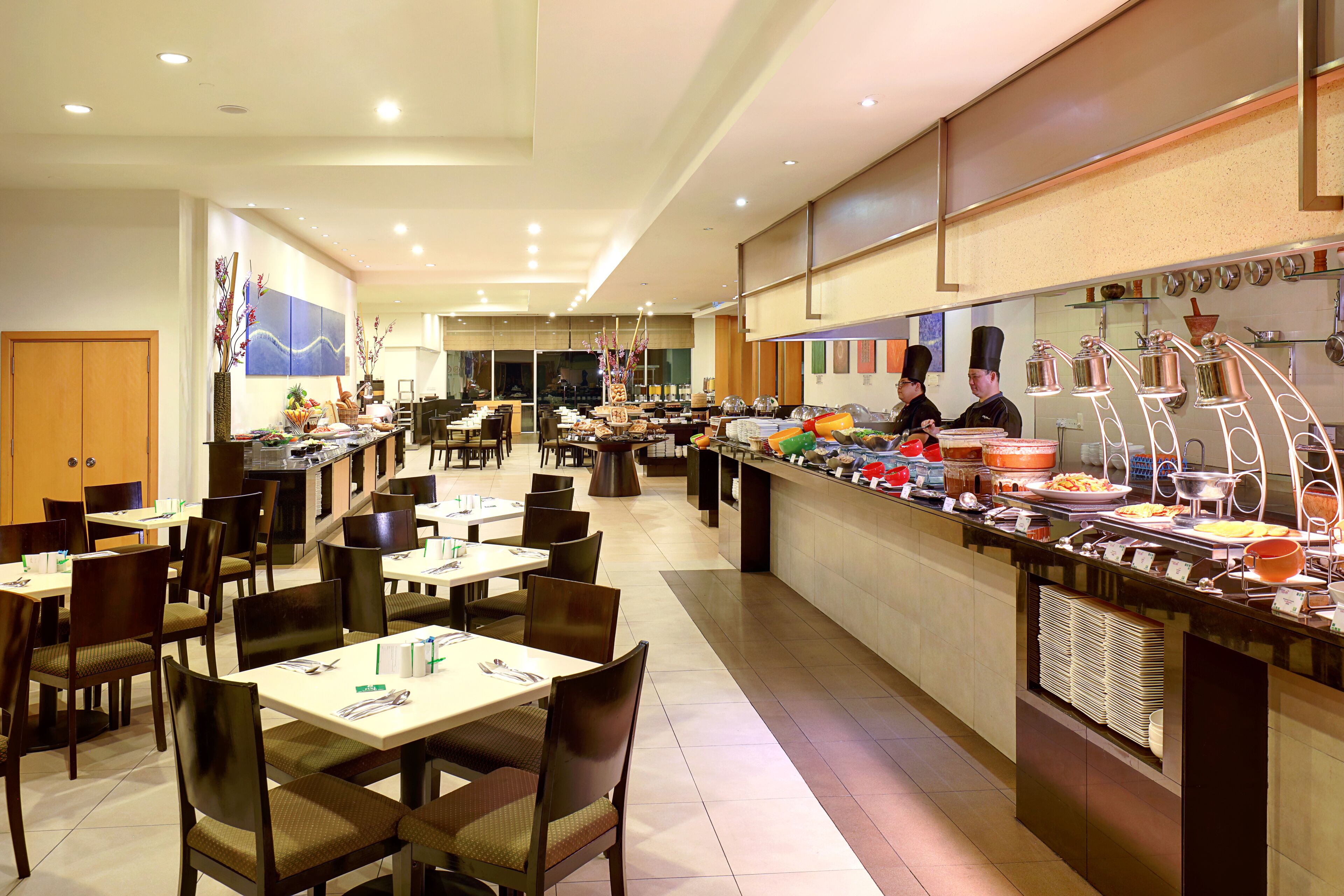 A buffet-style restaurant with multiple dining tables and chairs, a counter with various food items, and a kitchen area in the background.