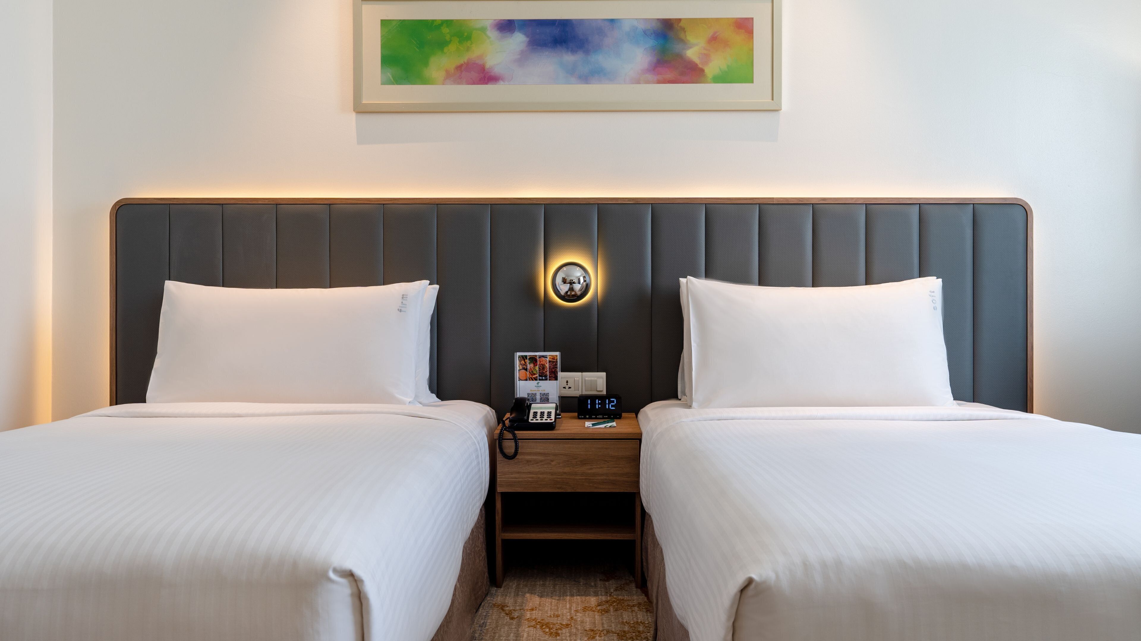 Habitación de hotel con dos camas, una mesita de noche con un teléfono y un reloj digital, y un cuadro abstracto enmarcado en la pared.