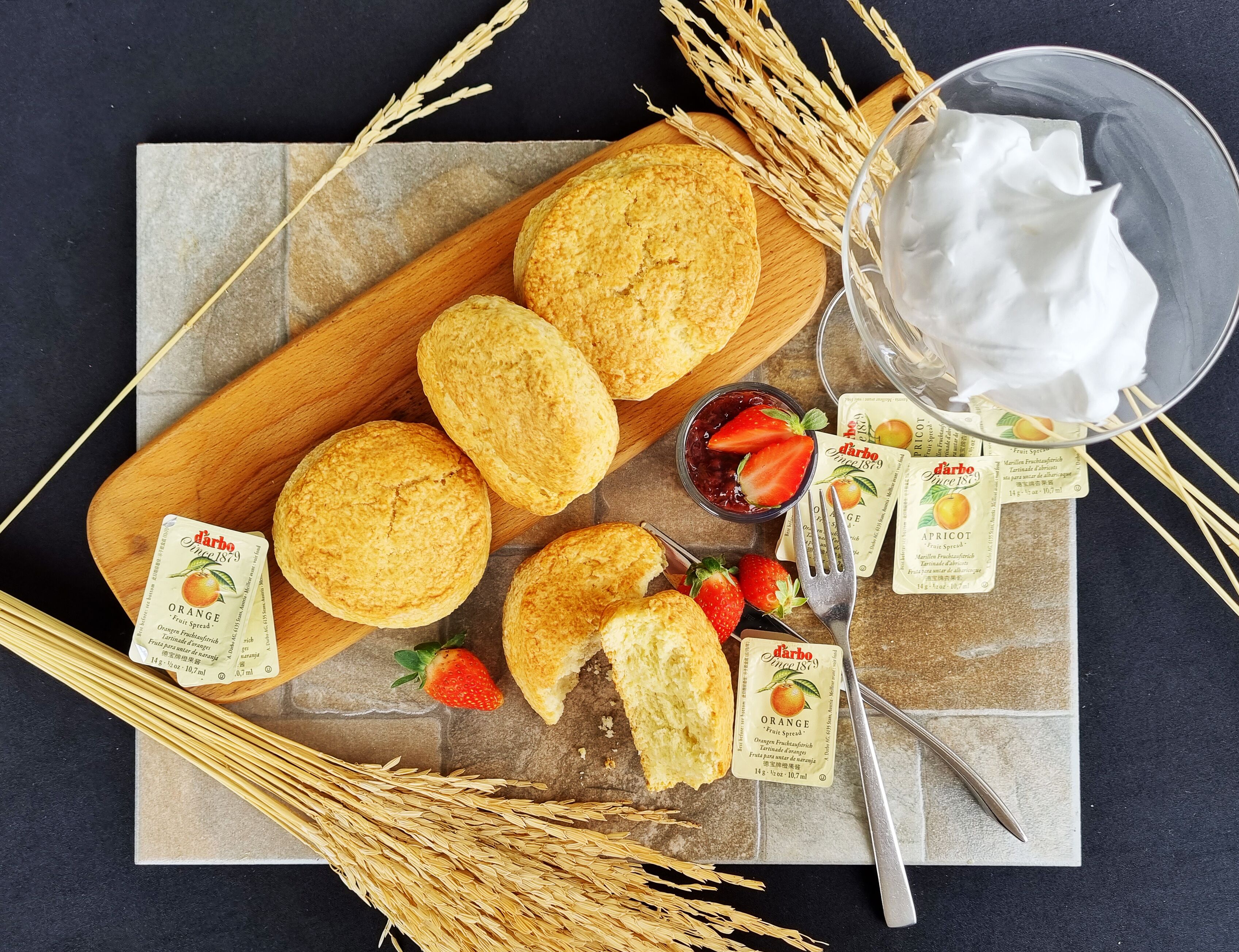 A wooden board with biscuits, a jar of jam, and a bowl of whipped cream.