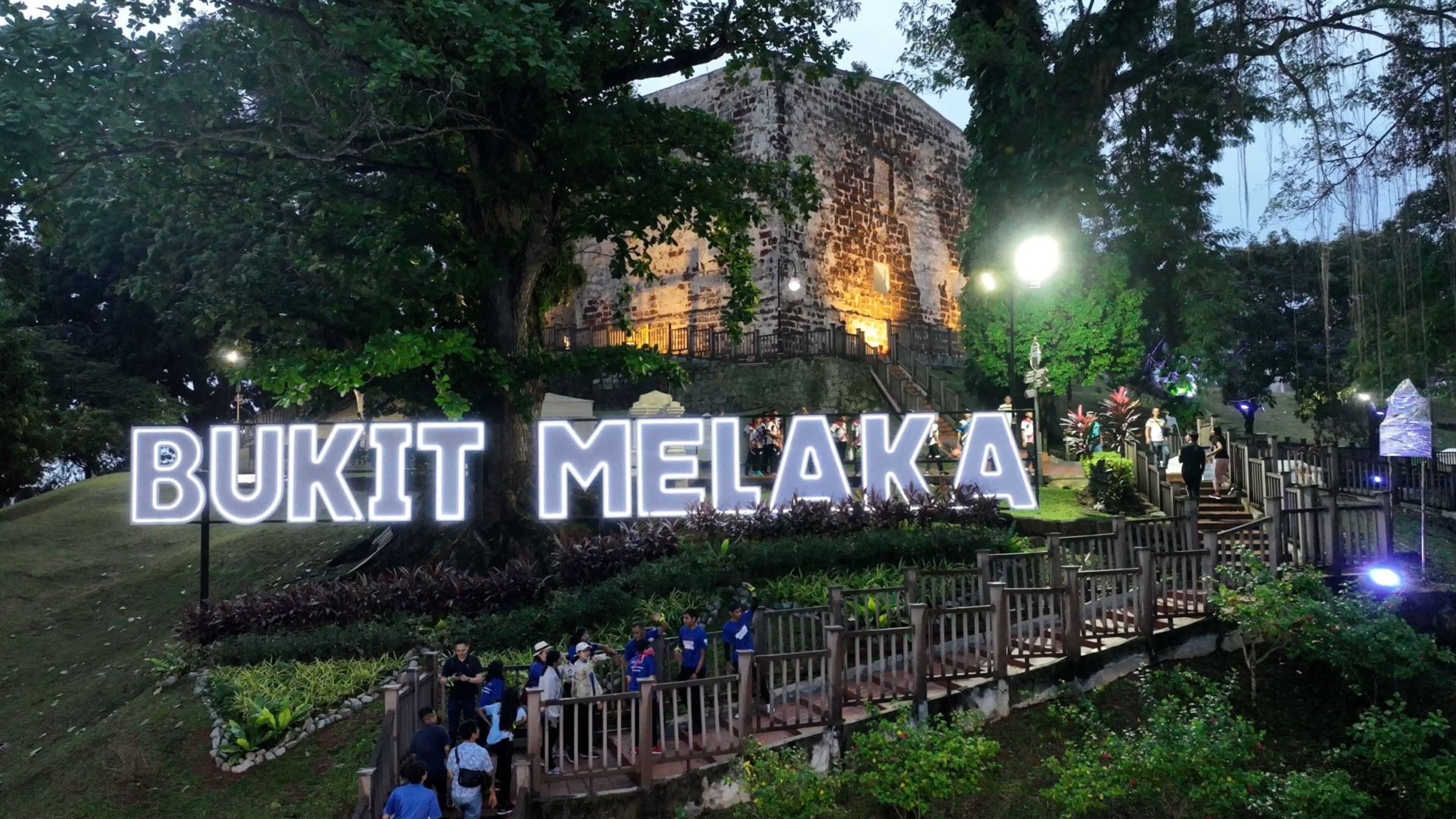 Un edificio histórico con un letrero que dice "BUKIT MELAKA", rodeado de follaje y un pasillo de madera.
