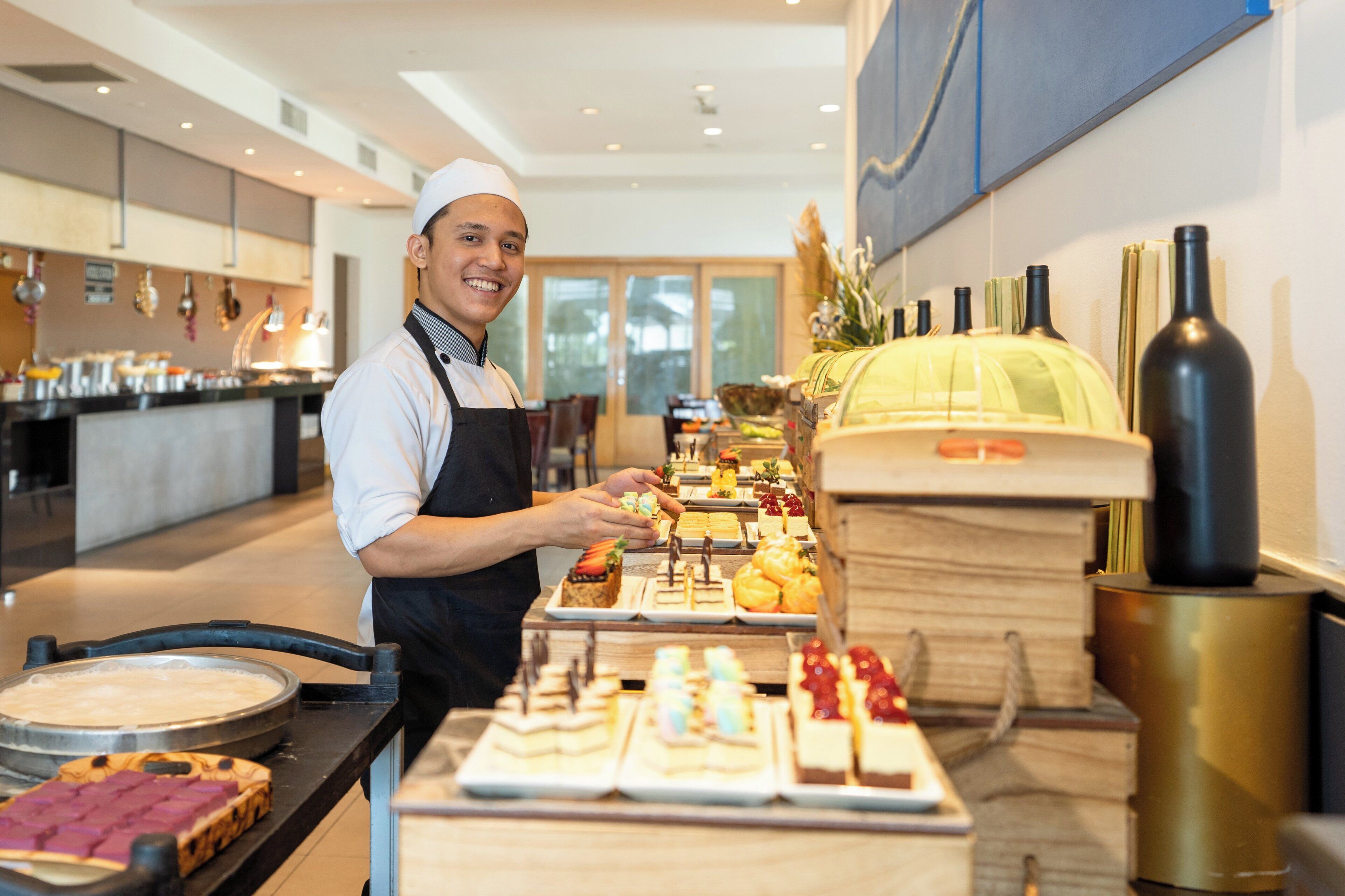 A person serving food at a buffet with various dishes and a wine bottle on the counter.