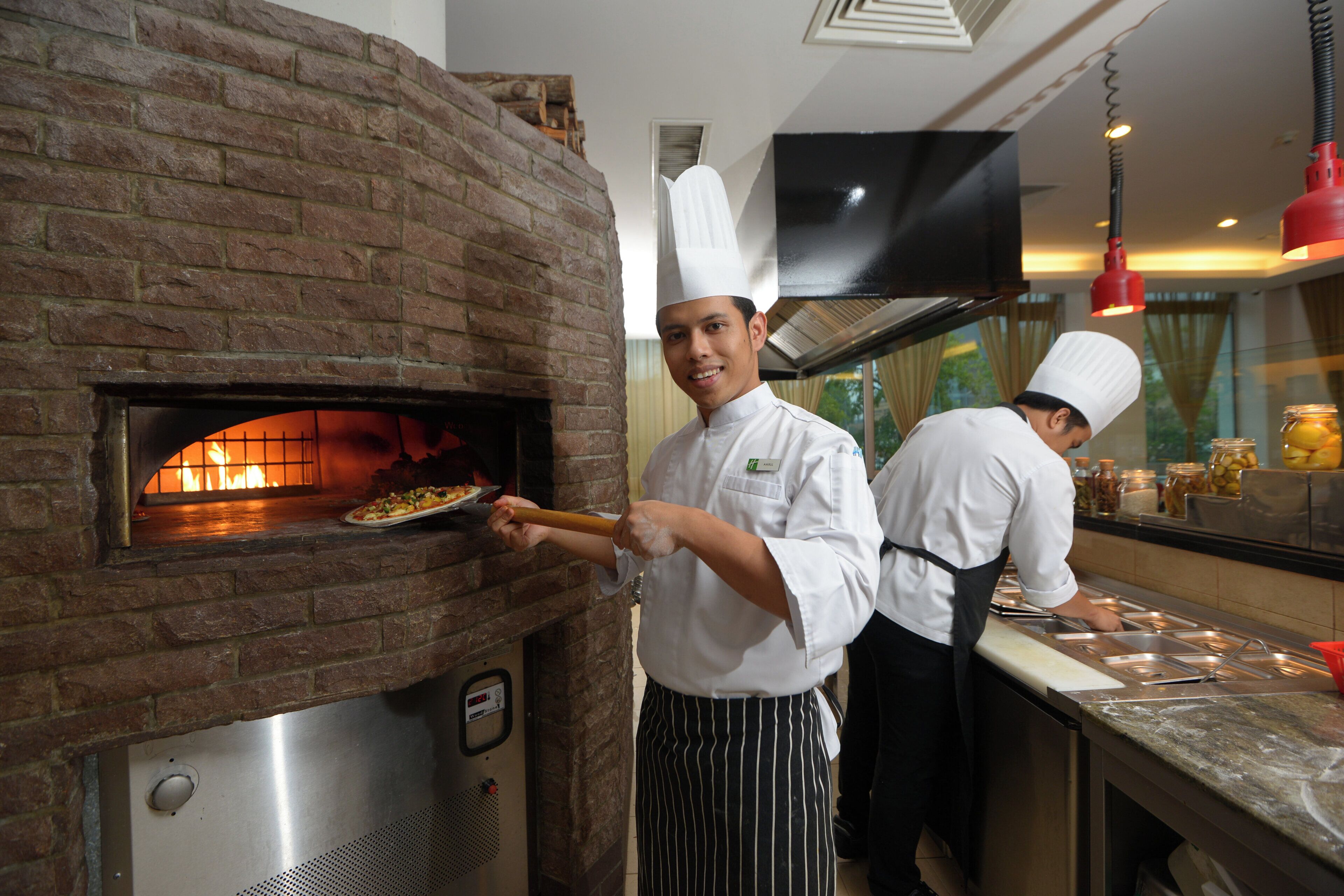 Un chef usa un horno de ladrillo para hornear pizza, mientras otro chef prepara comida al fondo.