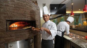 Un chef usa un horno de ladrillo para hornear pizza, mientras otro chef prepara comida al fondo.