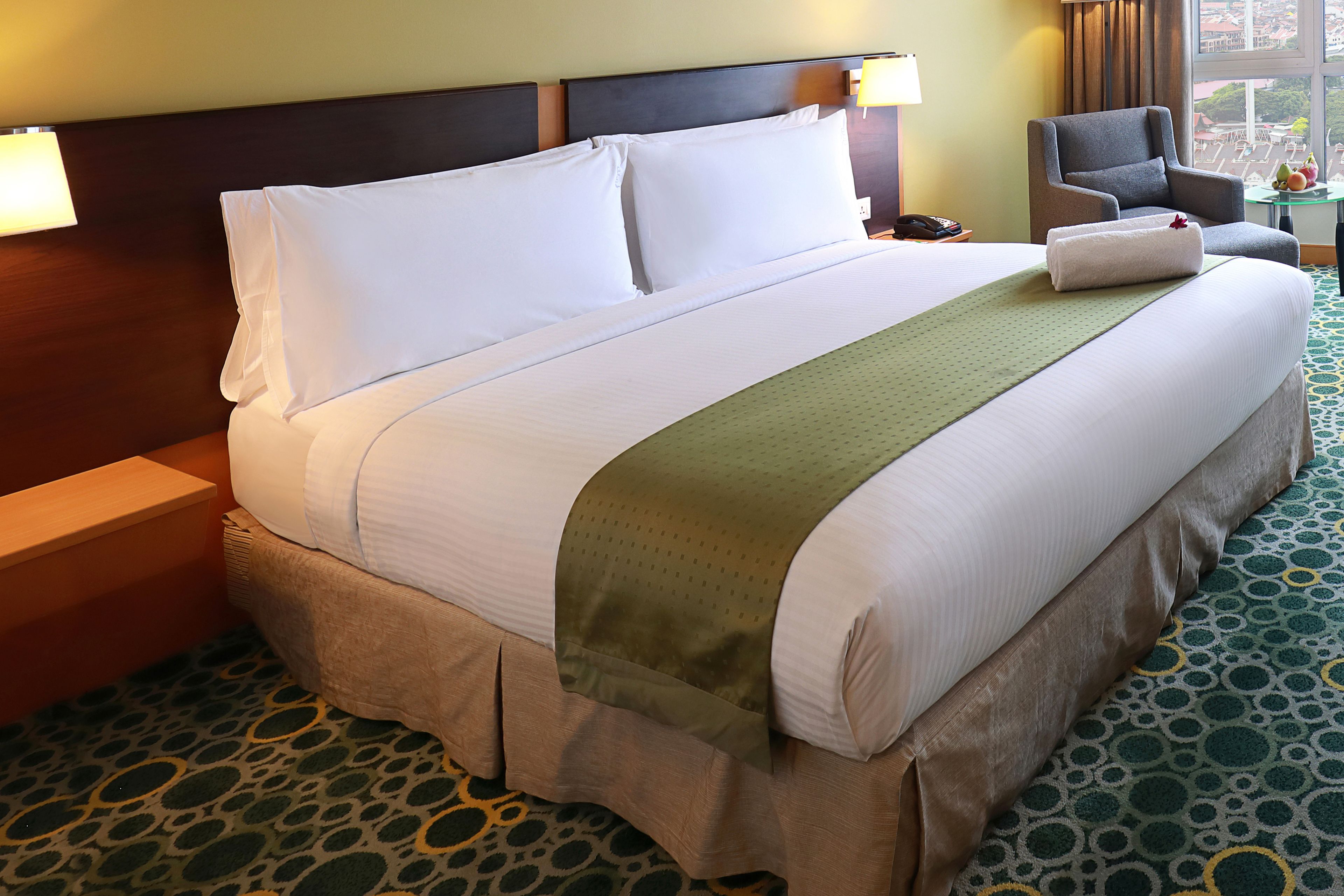 A hotel room with a large bed, a nightstand, a chair, a small table with fruit, and a view of the city through the window.