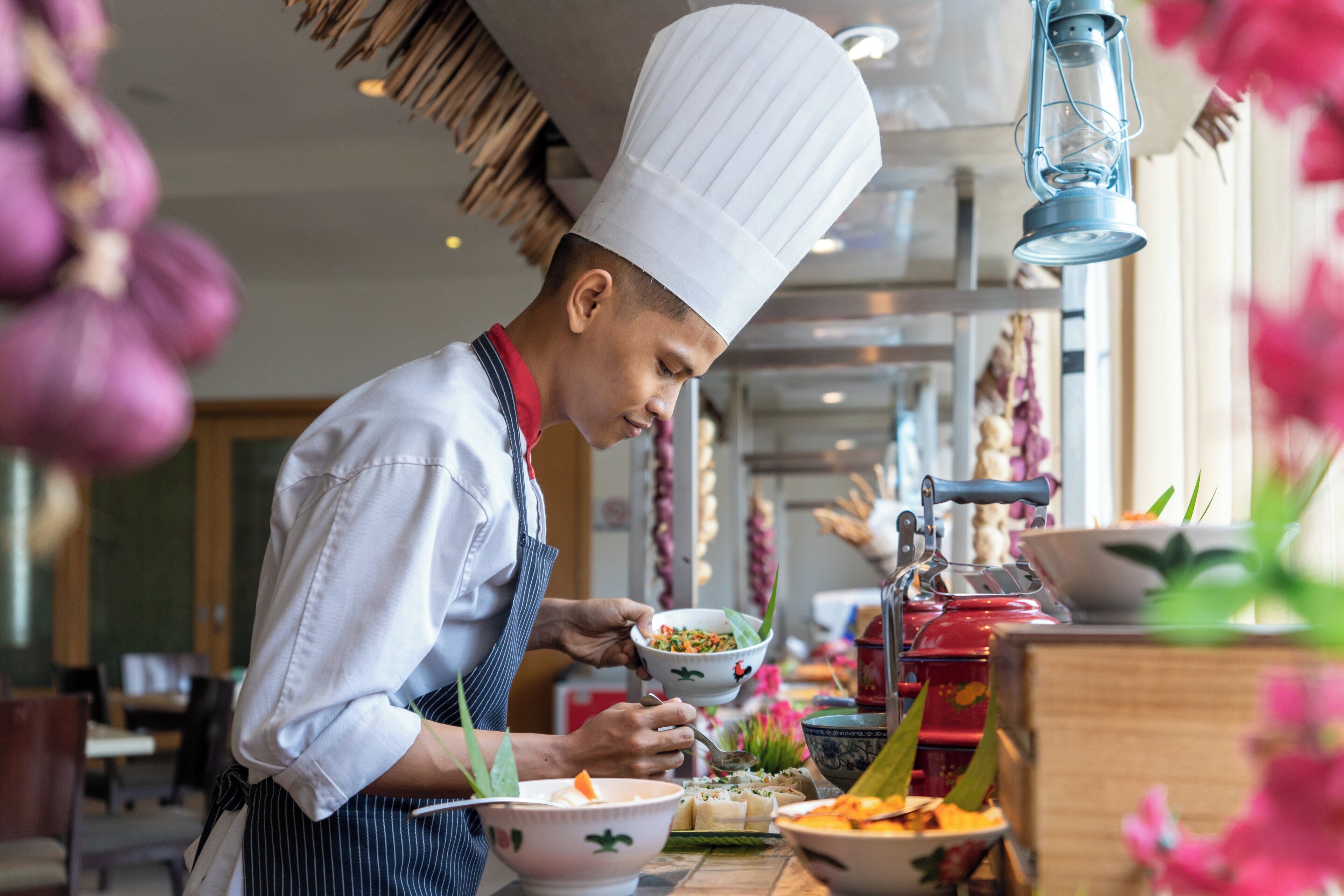 A chef in a white hat and striped apron preparing food in a kitchen with a traditional stove and various dishes on the counter.