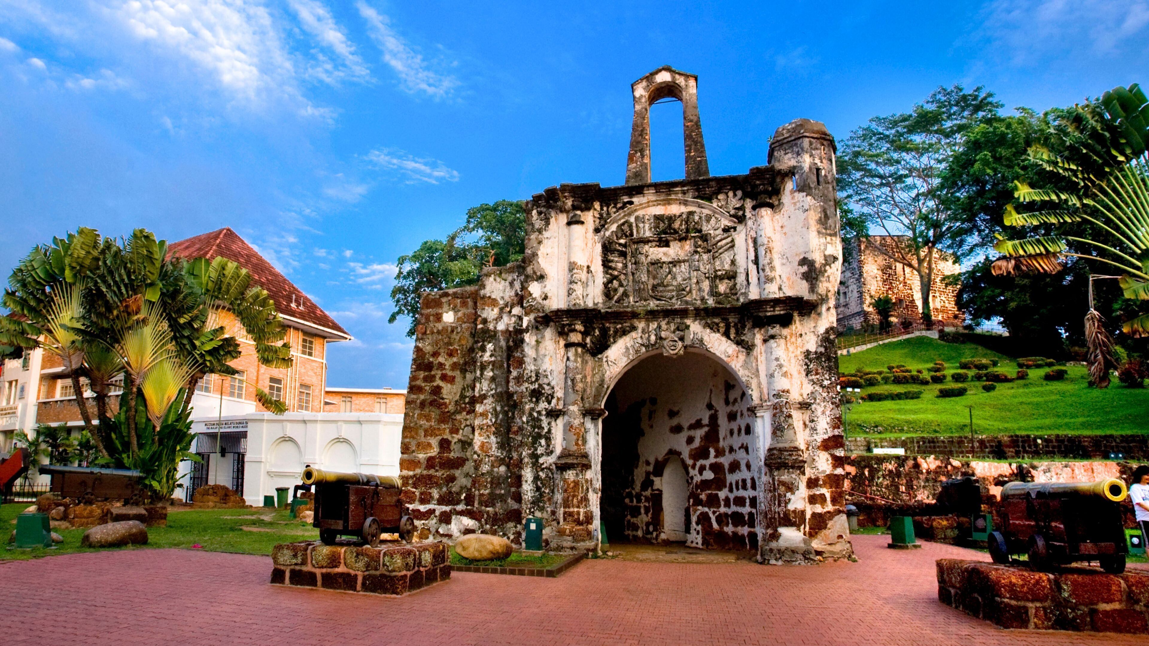 Un antiguo portón de piedra con un reloj, rodeado de palmeras y un edificio moderno al fondo.