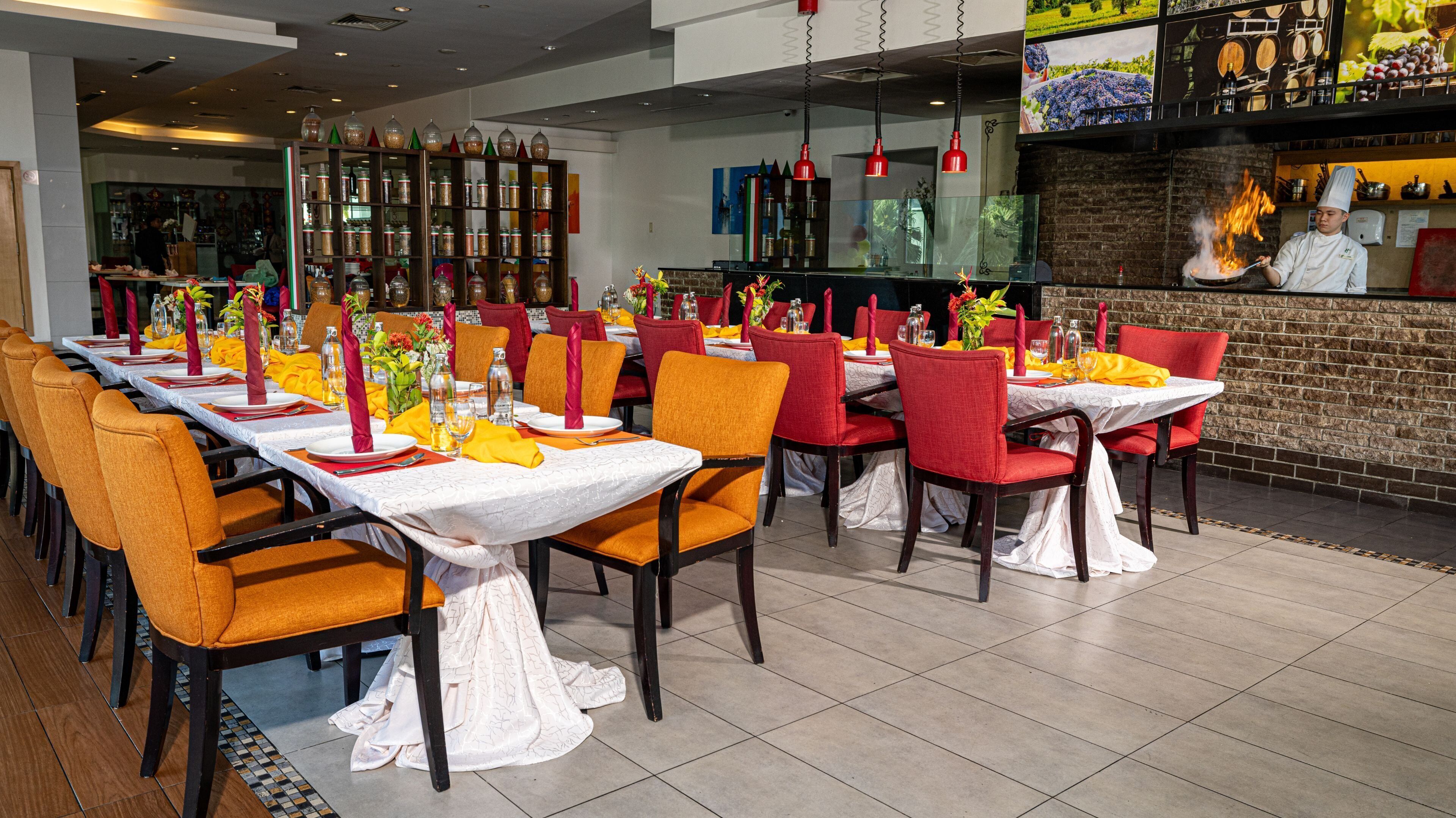Un restaurant avec des tables dressées pour le service, un bar où un chef prépare les plats, et un mur décoratif orné d’images.