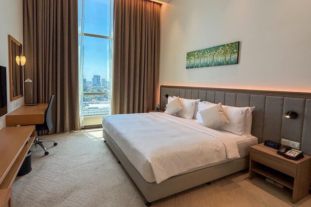 Ein Hotelzimmer mit einem großen Bett, einem Schreibtisch mit Fernseher, einem Bild an der Wand und Blick auf die Stadt.