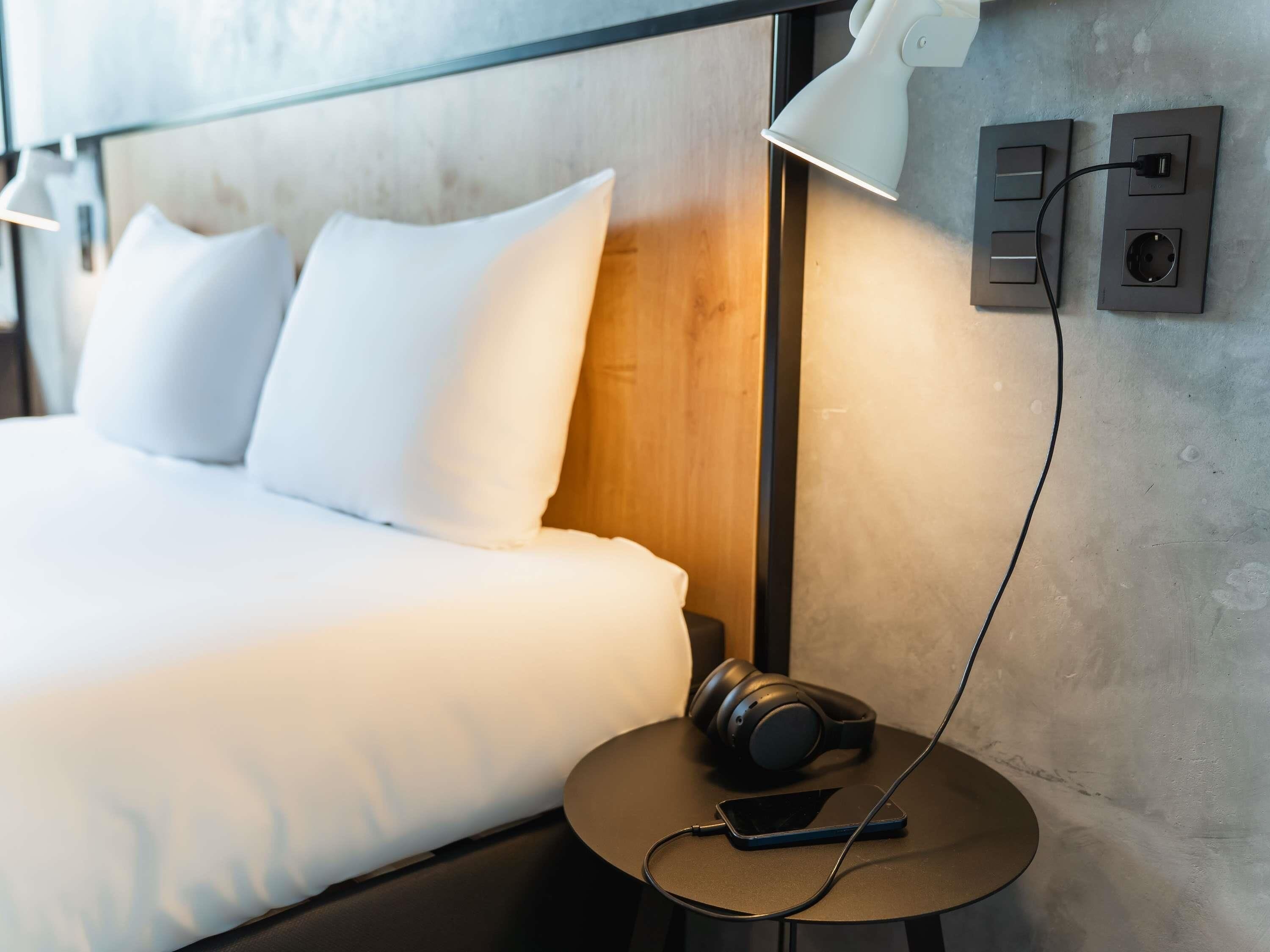A hotel room with a bed, pillows, a bedside table with headphones and a smartphone, a lamp, and a wall socket.