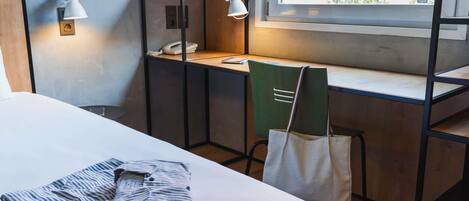 A hotel room with a bed, desk, and chair. There are two wall-mounted lamps, a window with a view of greenery, and a telephone on the desk.