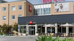 Et hotel med et tydeligt 'ibis HOTEL' skilt, udendørs siddepladser med parasoller, og et skilt der siger 'WELCOME INTO MY WORLD'.